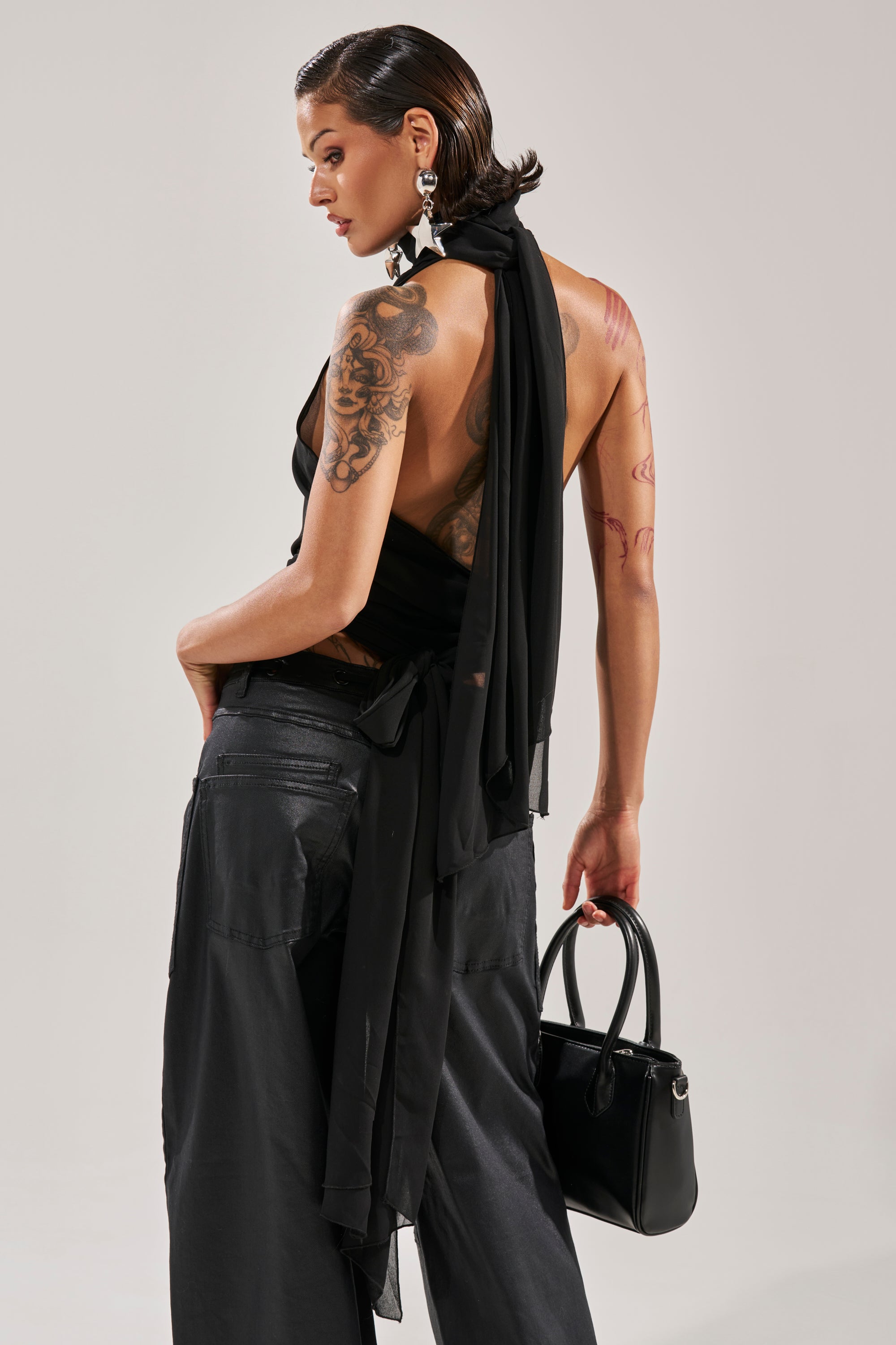 A person with short, slicked-back hair, tattooed arms, and geometric earrings wears the black GOT IT FROM YOU BLOUSE with matching pants and a black handbag, posing against a plain light background. 5
