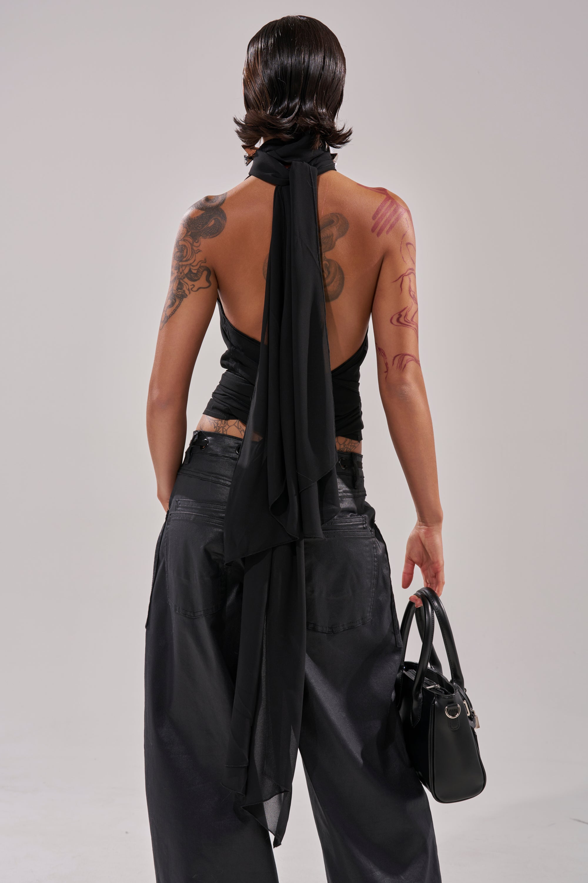 A tattooed person, seen from the back against a plain light background, wears the GOT IT FROM YOU BLOUSE—a black halter wrap top with a long scarf—paired with loose black pants and holding a black handbag. 6
