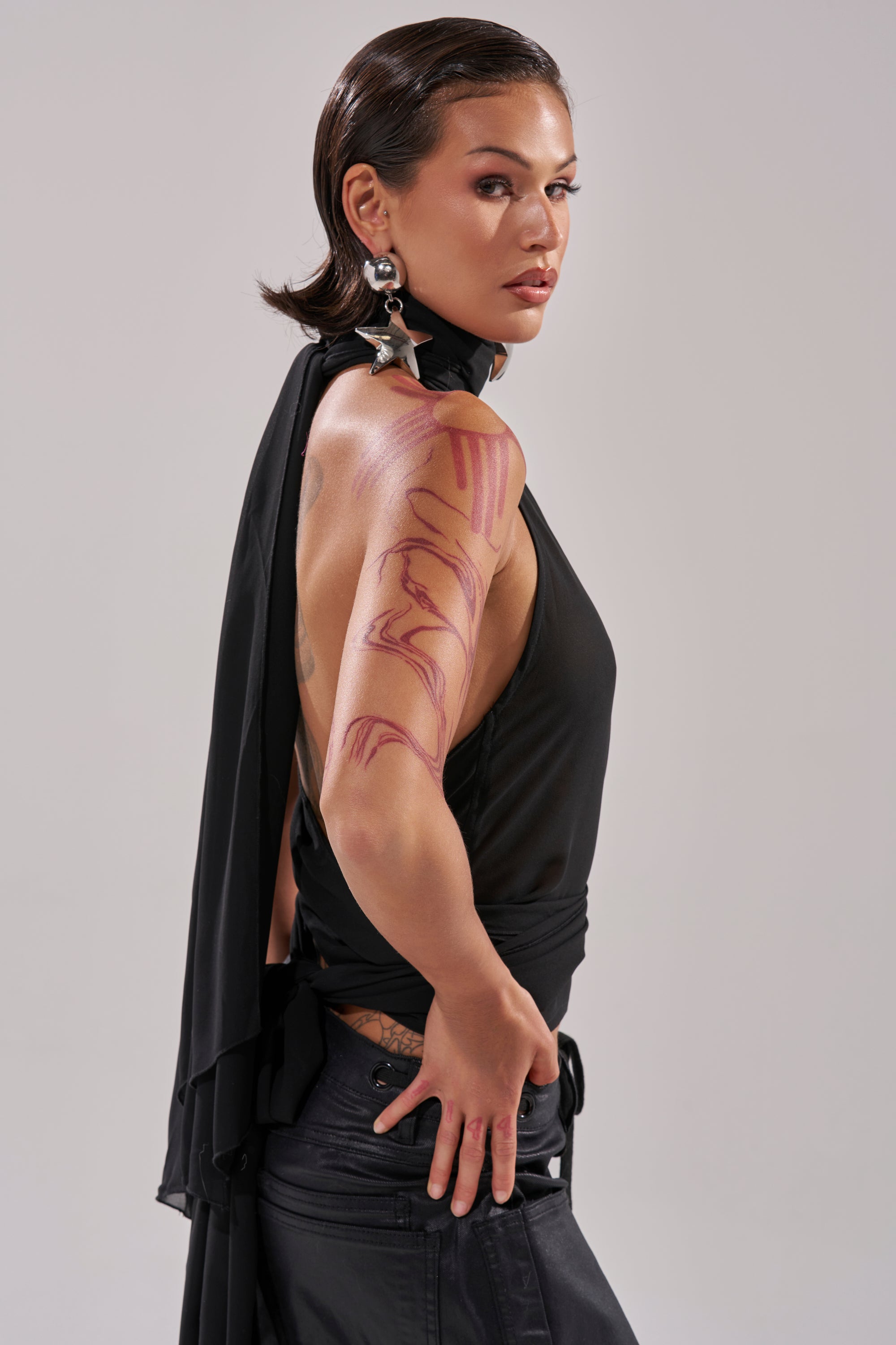 A person with short, slicked-back hair poses confidently over their shoulder in the sleek GOT IT FROM YOU BLOUSE and dark pants, accessorized with large star-shaped earrings and displaying an abstract pink tattoo on their arm. 7