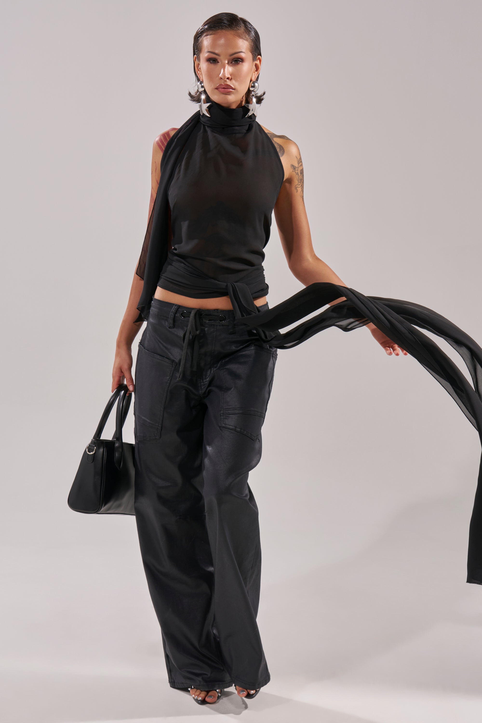 A person poses confidently in the GOT IT FROM YOU BLOUSE—a black halter wrap top—paired with loose black cargo pants, heels, a black handbag, and earrings against a plain light background. 8