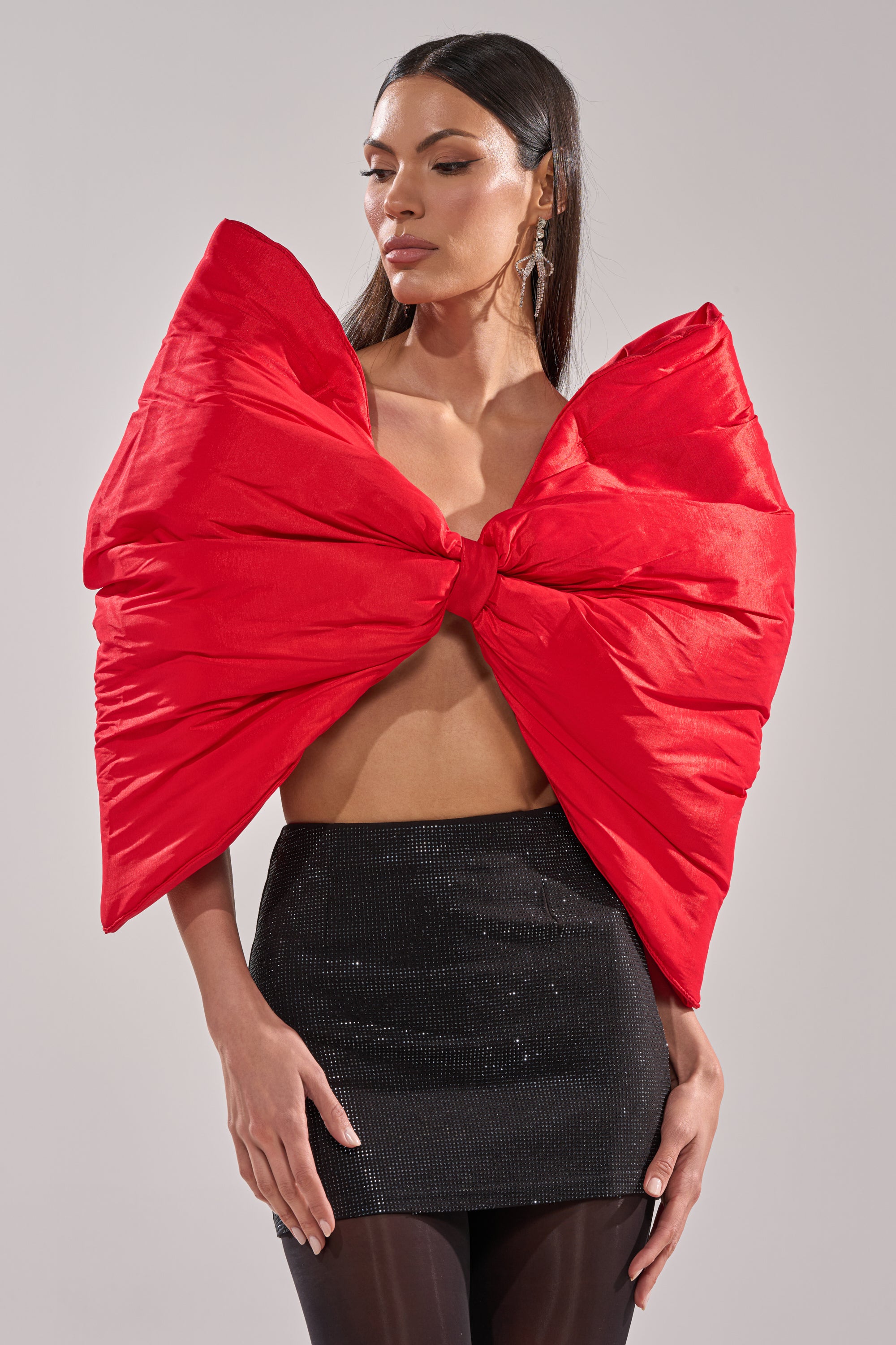 A woman with long, straight dark hair stands against a neutral background wearing the EXTRA BIG BOW TOP in bold red satin, styled with a short black textured skirt and sheer black tights, looking slightly downward.