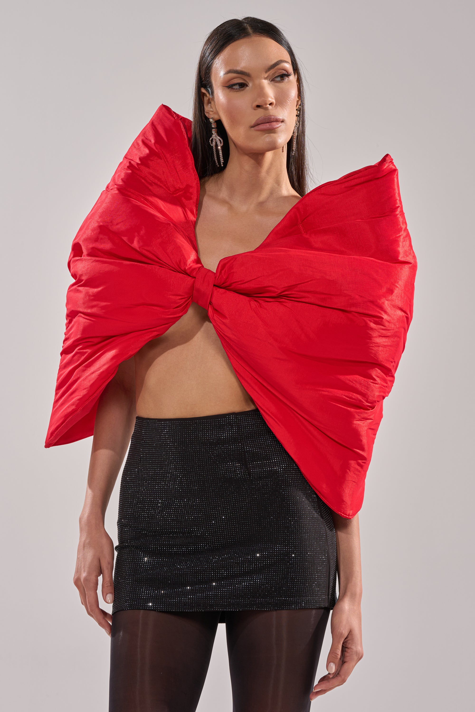 A woman models the EXTRA BIG BOW TOP in a satin-like fabric, styled with a short black skirt and sheer black tights, against a plain light backdrop.