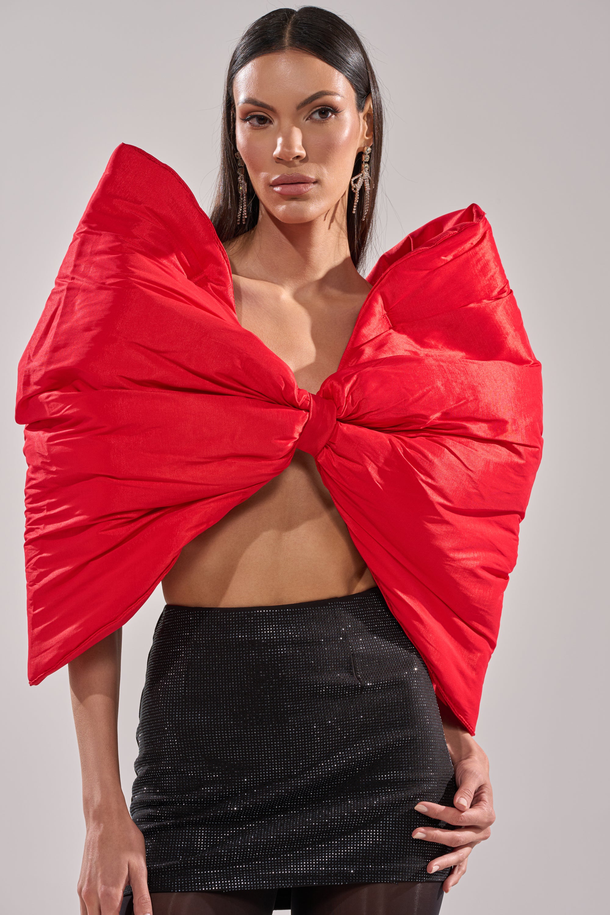 A woman models the EXTRA BIG BOW TOP in bold red satin and a short black skirt, standing against a plain background with straight dark hair, looking confidently at the camera.