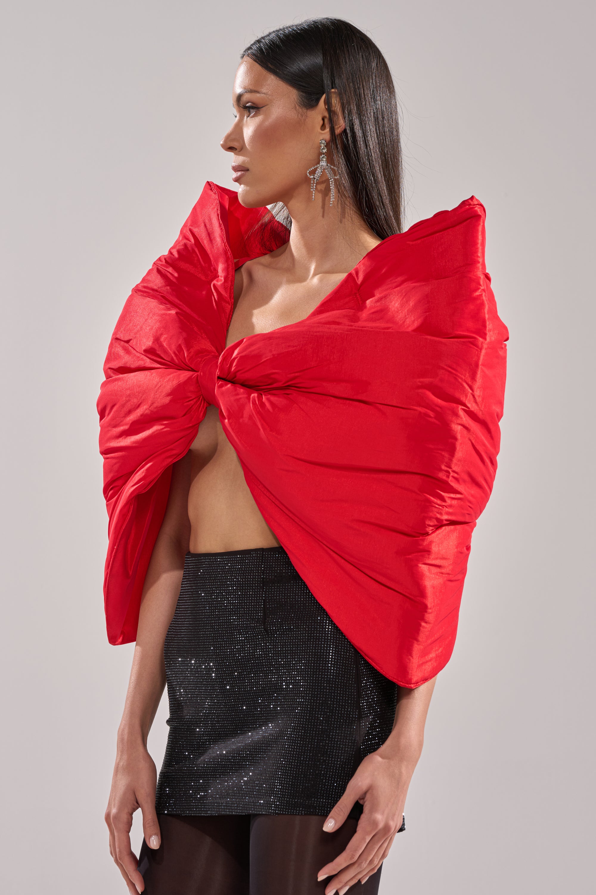 A woman with straight dark hair wears the EXTRA BIG BOW TOP in dramatic satin, a short black sequined skirt, sheer black tights, and dangling earrings, standing against a plain light background.