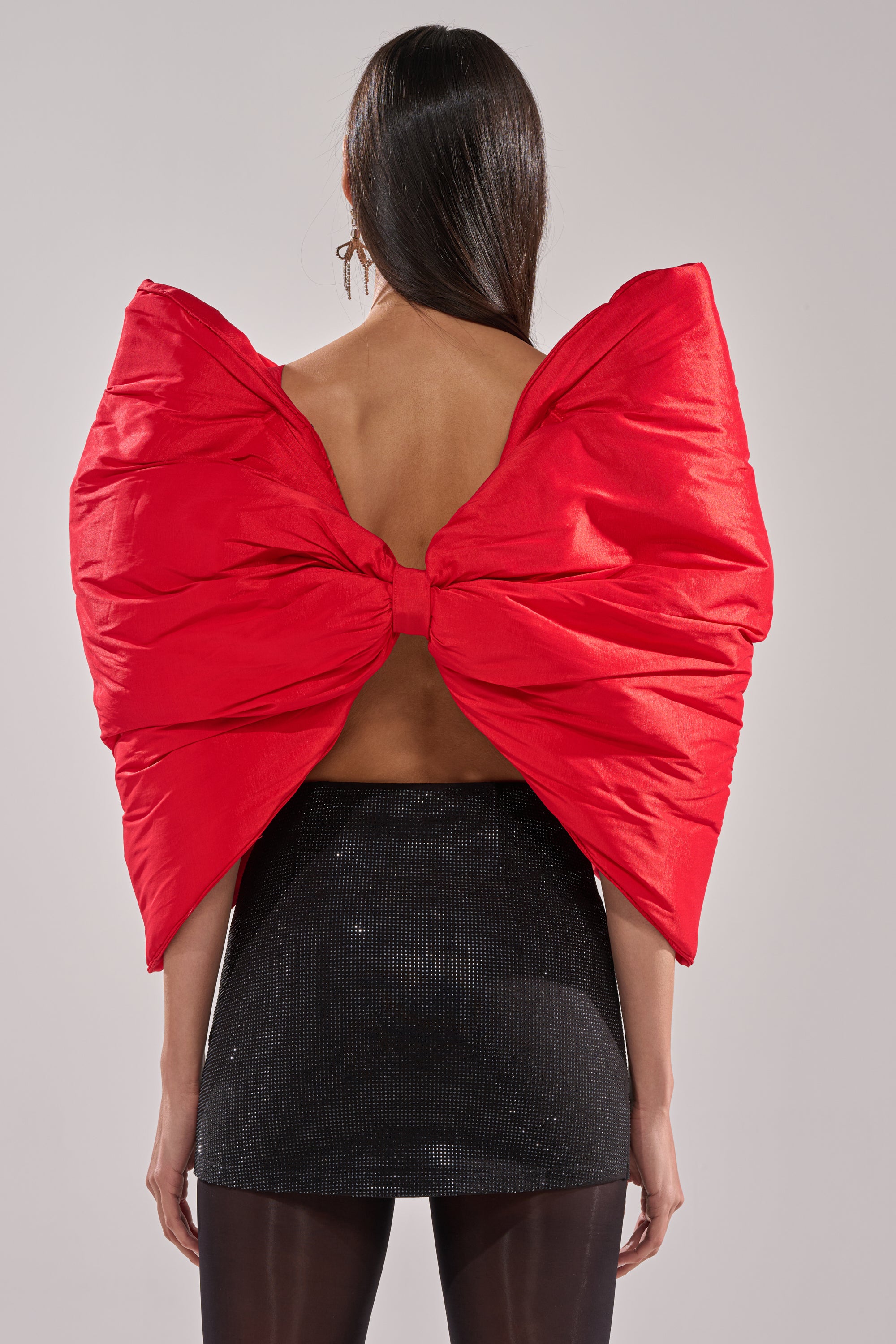 A woman with long dark hair, photographed from behind against a plain background, wears the EXTRA BIG BOW TOP in bright red satin—featuring an open back—paired with a short black skirt and black tights.