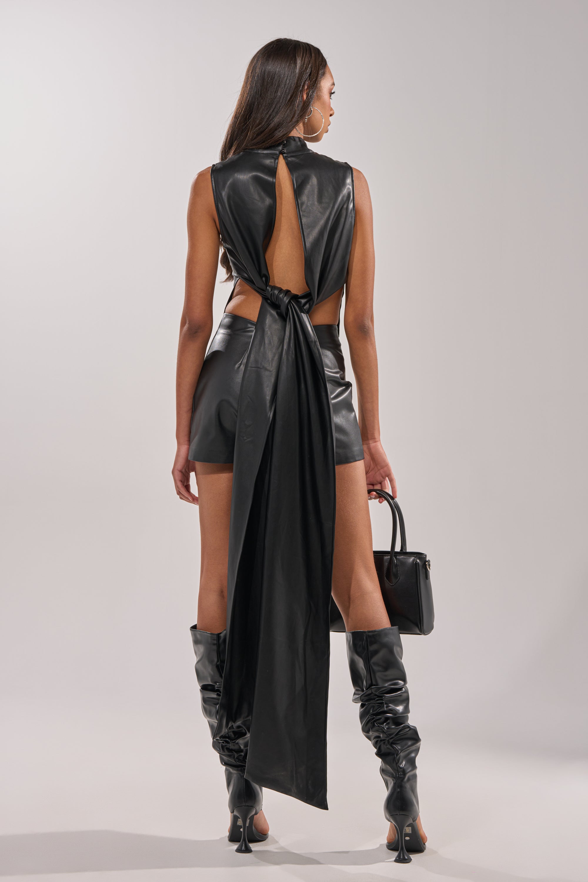 A person faces away, wearing the BABY GIRL FAUX LEATHER BLOUSE—a black sleeveless, open-back top with a long draped panel—paired with matching shorts, knee-high heeled boots, and a small handbag against a plain light background. 6