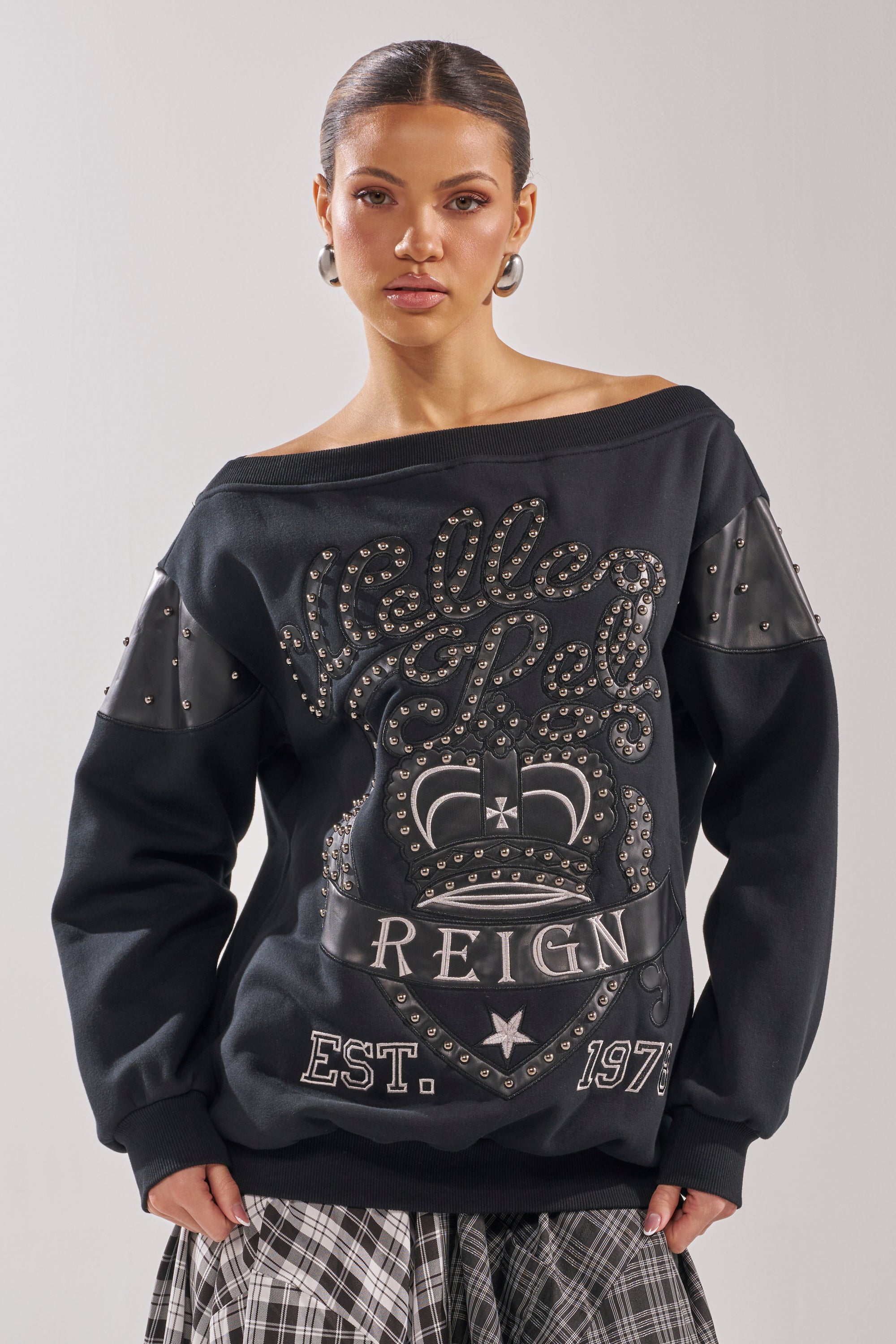 A woman wears the ROYAL OVERSIZED SWEATSHIRT, an off-the-shoulder pullover with studded lettering and a crown graphic, paired with a black-and-white plaid skirt. She stands facing forward against a plain background. 0