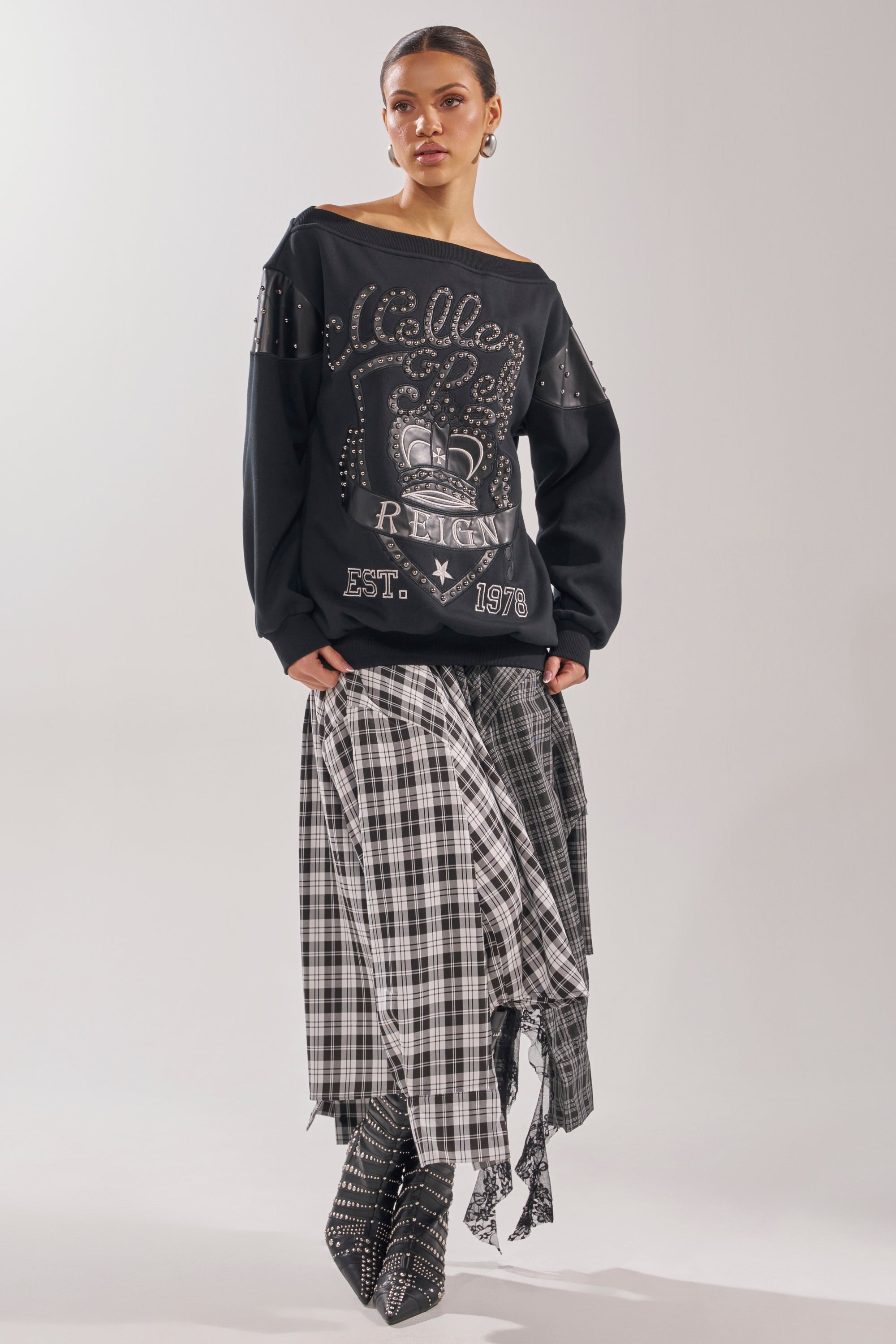 A woman with her hair pulled back and a neutral expression stands against a plain background wearing the ROYAL OVERSIZED SWEATSHIRT, styled with a layered black-and-white plaid skirt and pointed studded boots. 9