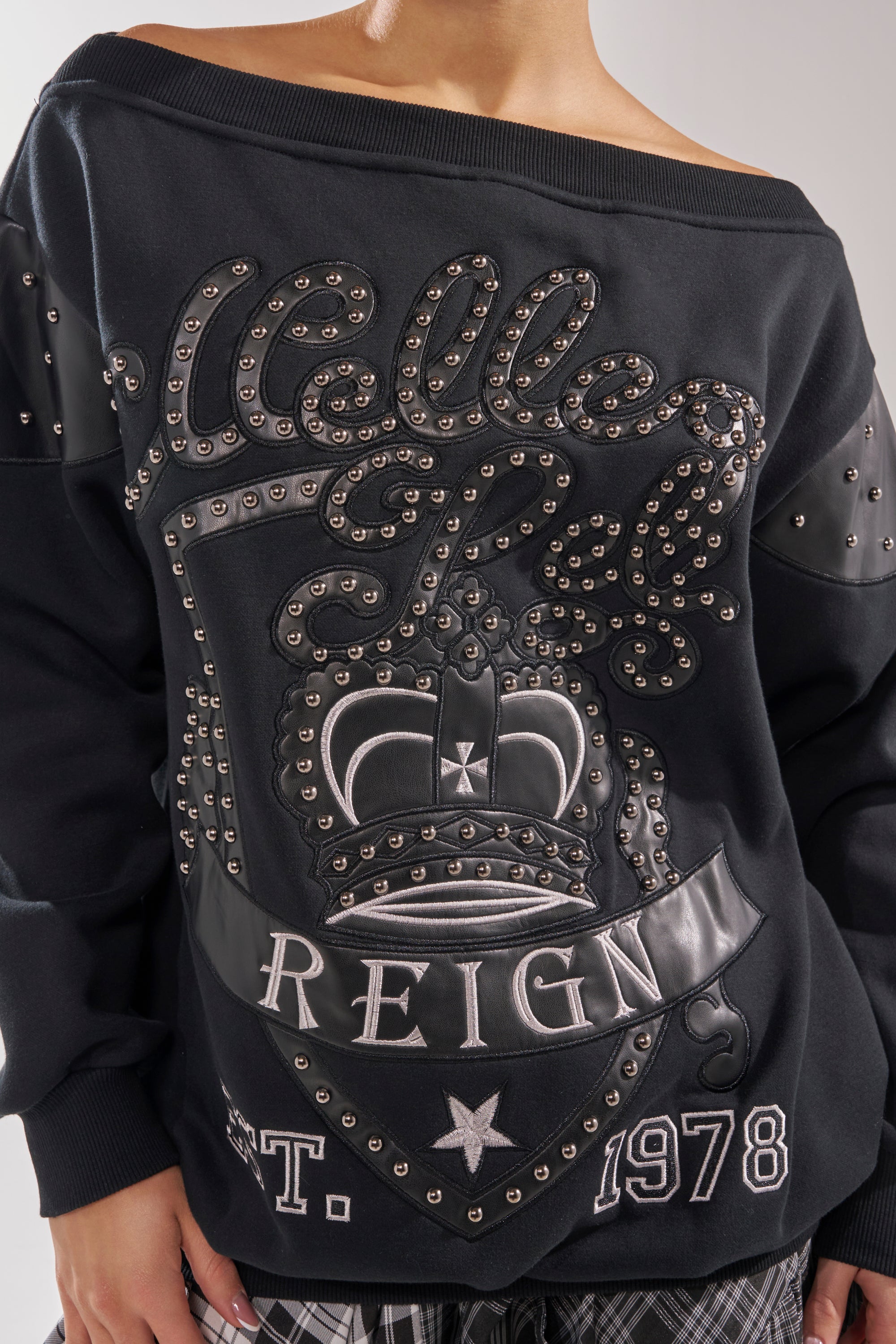 A person wears the ROYAL OVERSIZED SWEATSHIRT, an off-the-shoulder black streetwear piece with studded details, "Welles Reign," a crown graphic, and "EST. 1978," styled with plaid pants and their hand visible. 10
