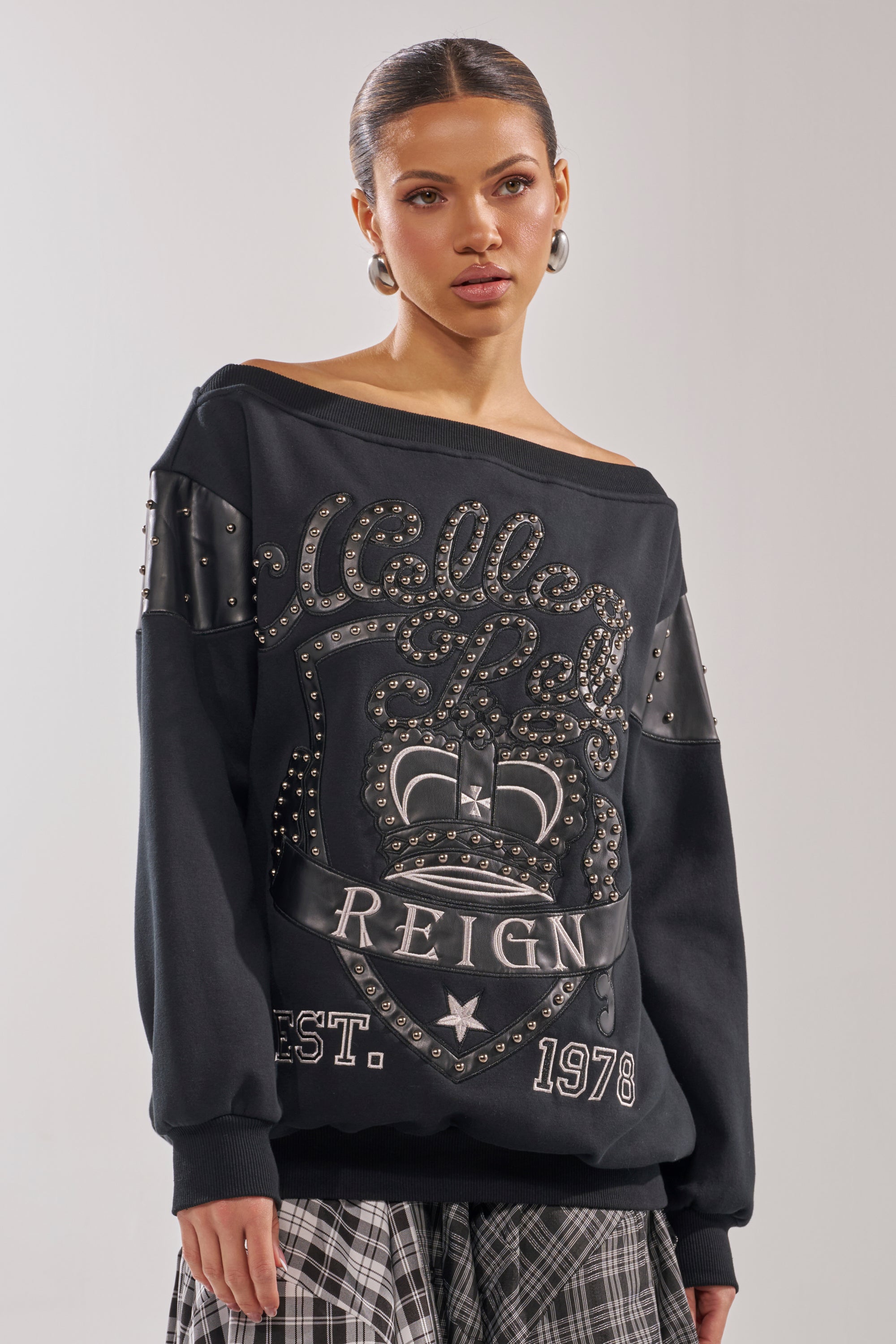 A woman models the ROYAL OVERSIZED SWEATSHIRT—black, off-the-shoulder, with metallic studs and bold graphic text—paired with black-and-white plaid pants against a plain background. 1