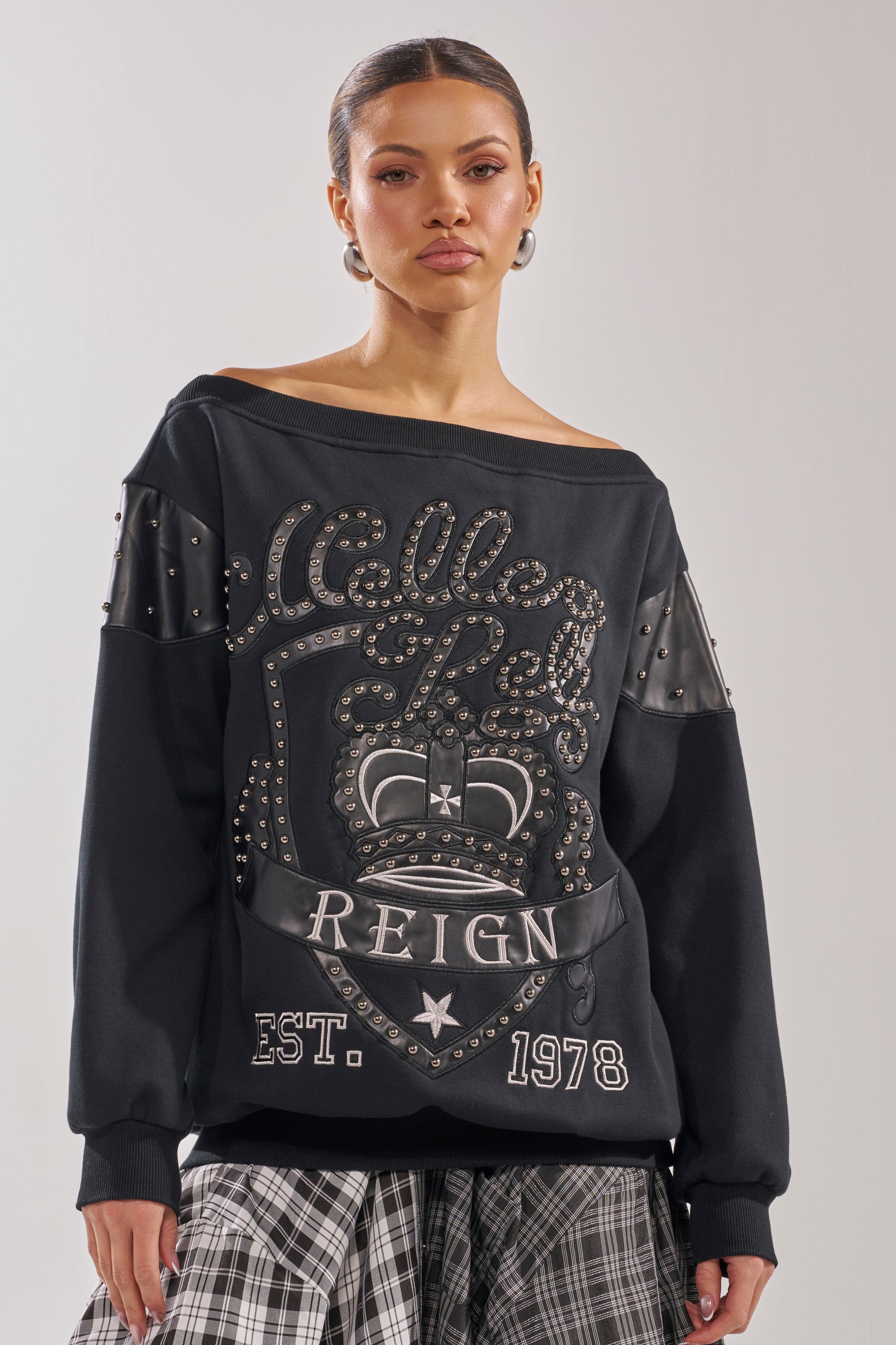 A woman models the ROYAL OVERSIZED SWEATSHIRT—an off-the-shoulder black top with studded embellishments and graphic text—paired with a black-and-white plaid skirt, highlighting modern streetwear style against a neutral backdrop. 2