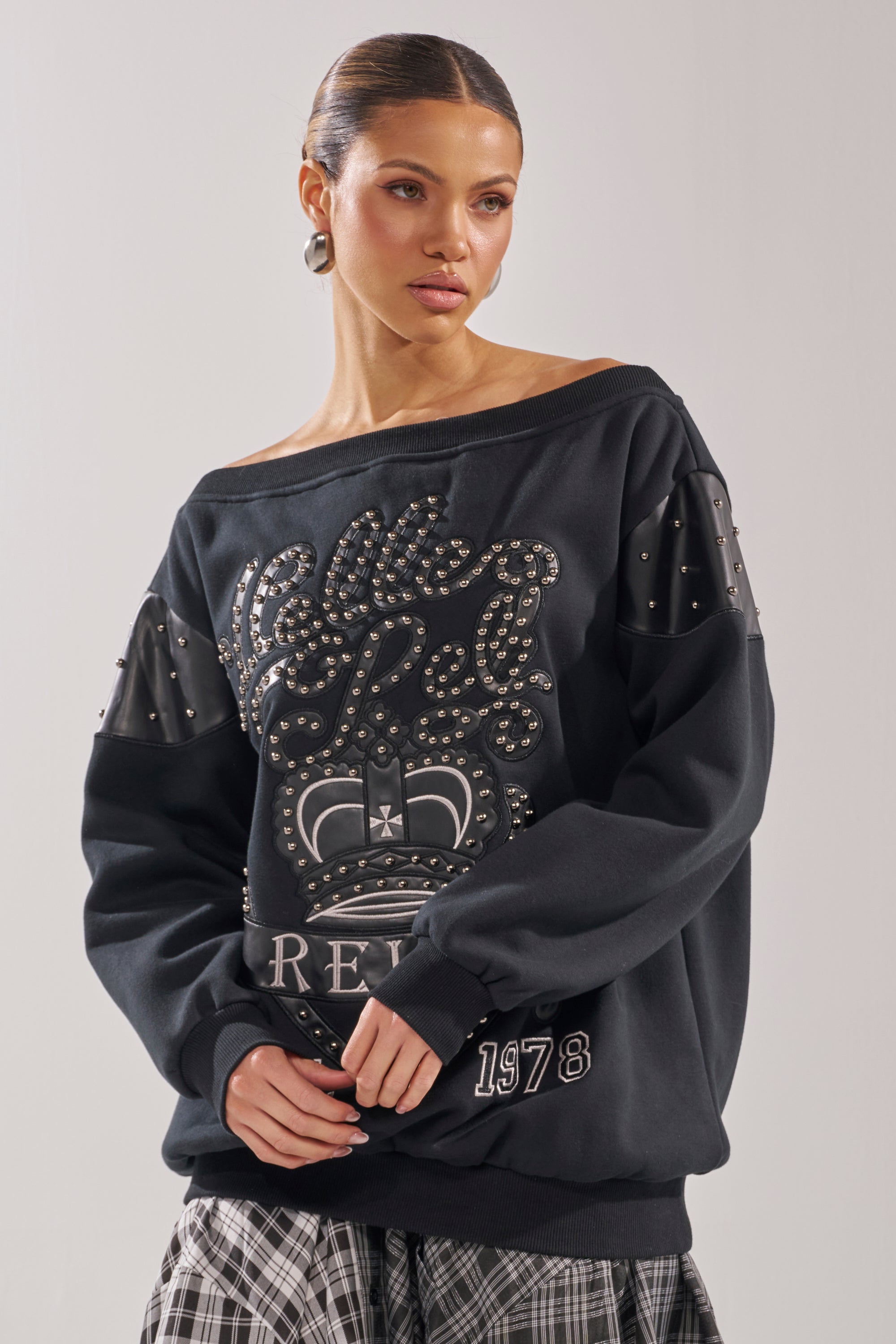 A woman wears the ROYAL OVERSIZED SWEATSHIRT—an off-the-shoulder black, streetwear-inspired top with metallic studs and a crown graphic—paired with a plaid skirt, hoop earrings, and her hair pulled back. 3