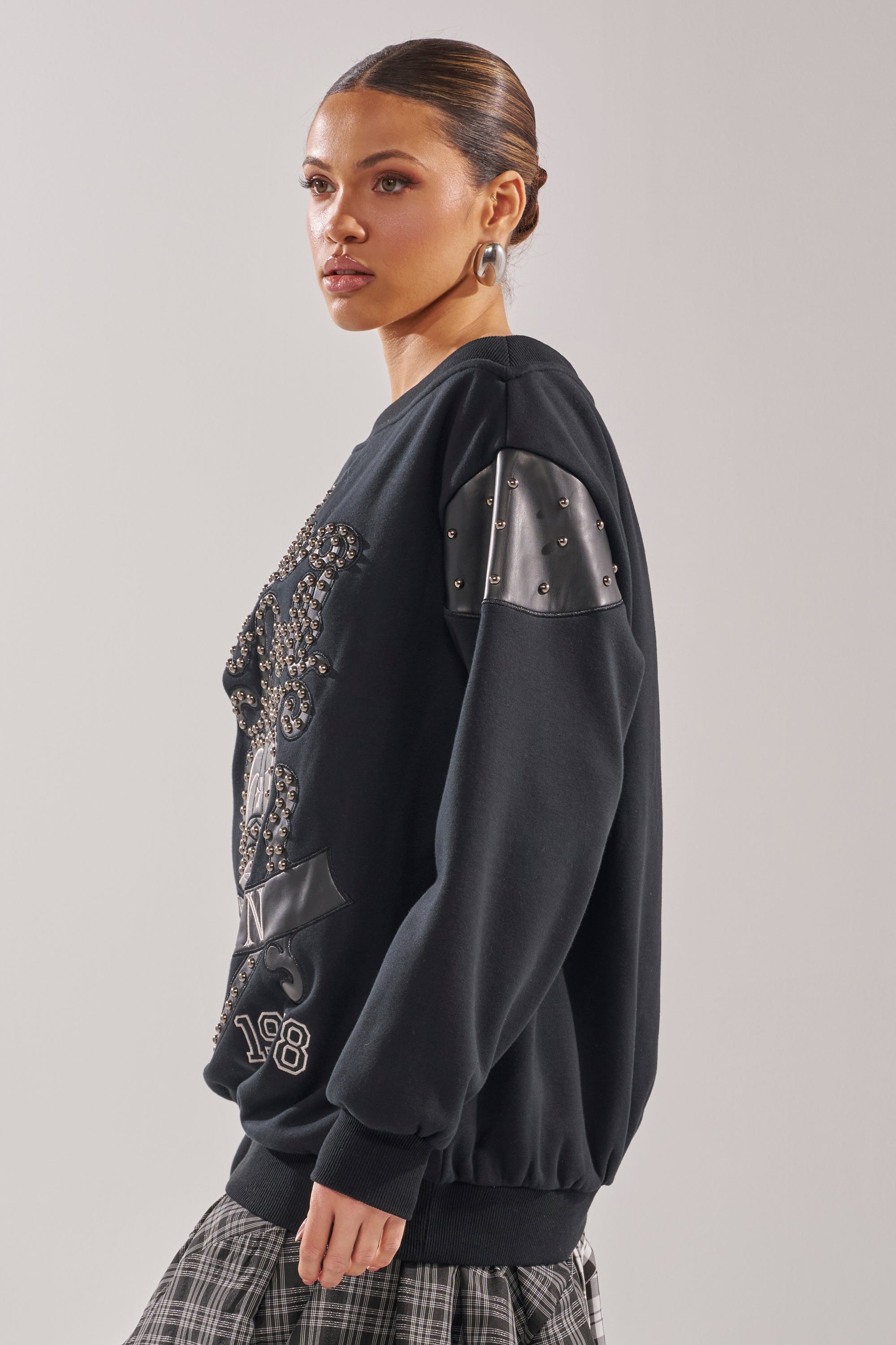 A woman with slicked-back hair models the ROYAL OVERSIZED SWEATSHIRT, featuring faux leather patches and studded details, paired with a plaid skirt against a plain light background. 4