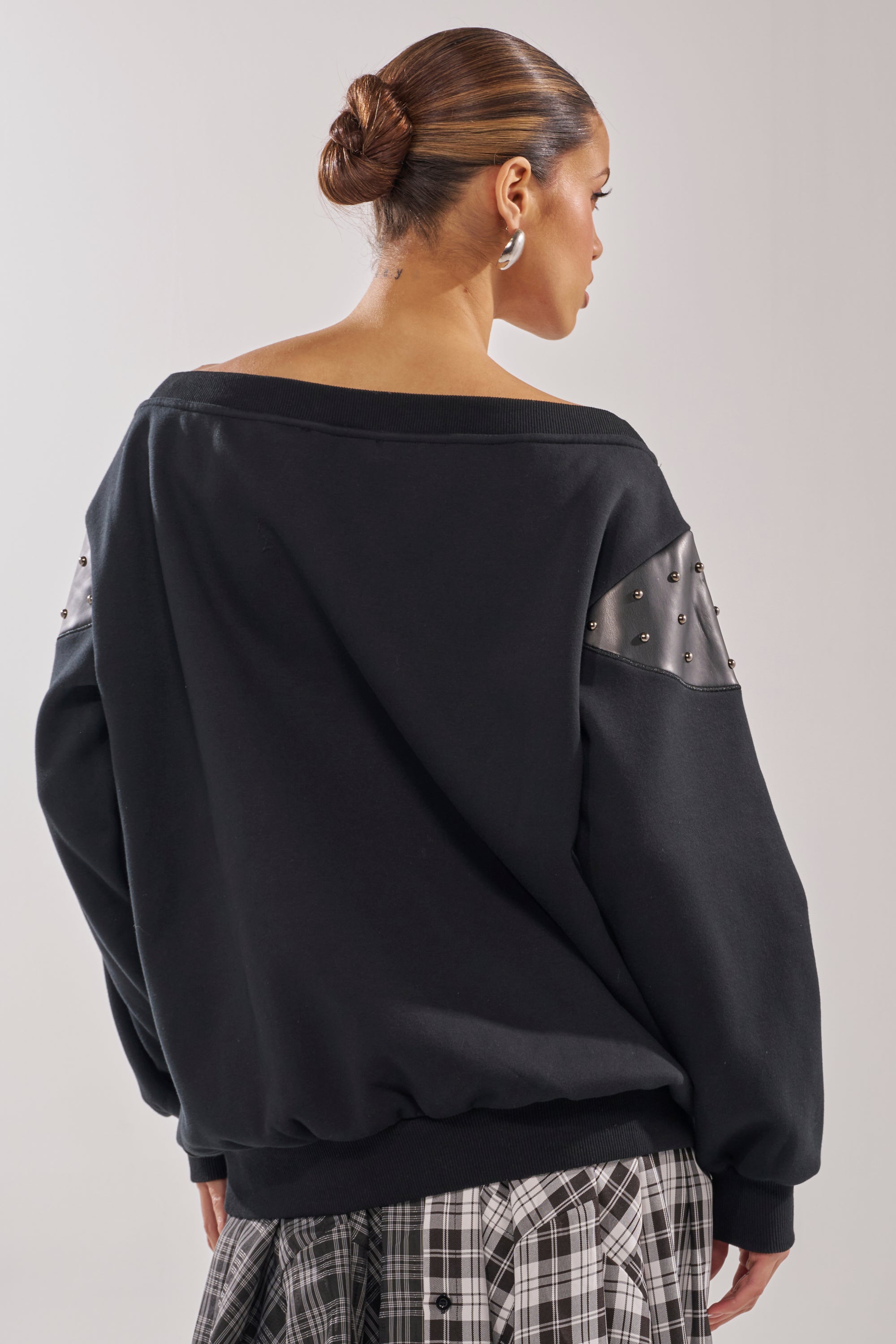 A woman with her hair in a sleek bun faces away, wearing the ROYAL OVERSIZED SWEATSHIRT featuring studded faux leather sleeve patches and pairing it with a black-and-white plaid skirt for a bold streetwear look. 5