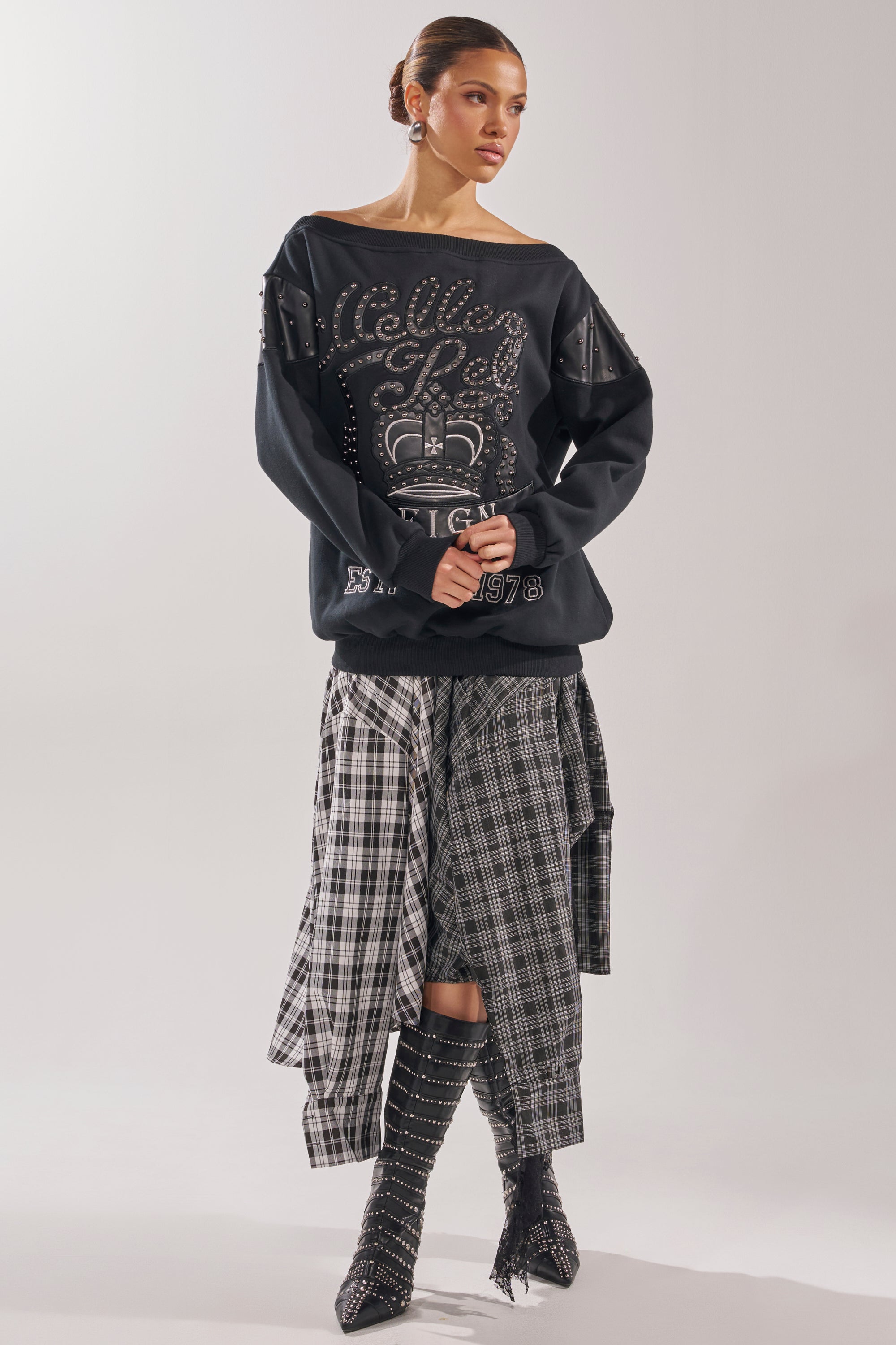 A woman models the ROYAL OVERSIZED SWEATSHIRT with an off-the-shoulder design, paired with a layered plaid skirt and black studded heeled boots, against a simple light background. 6