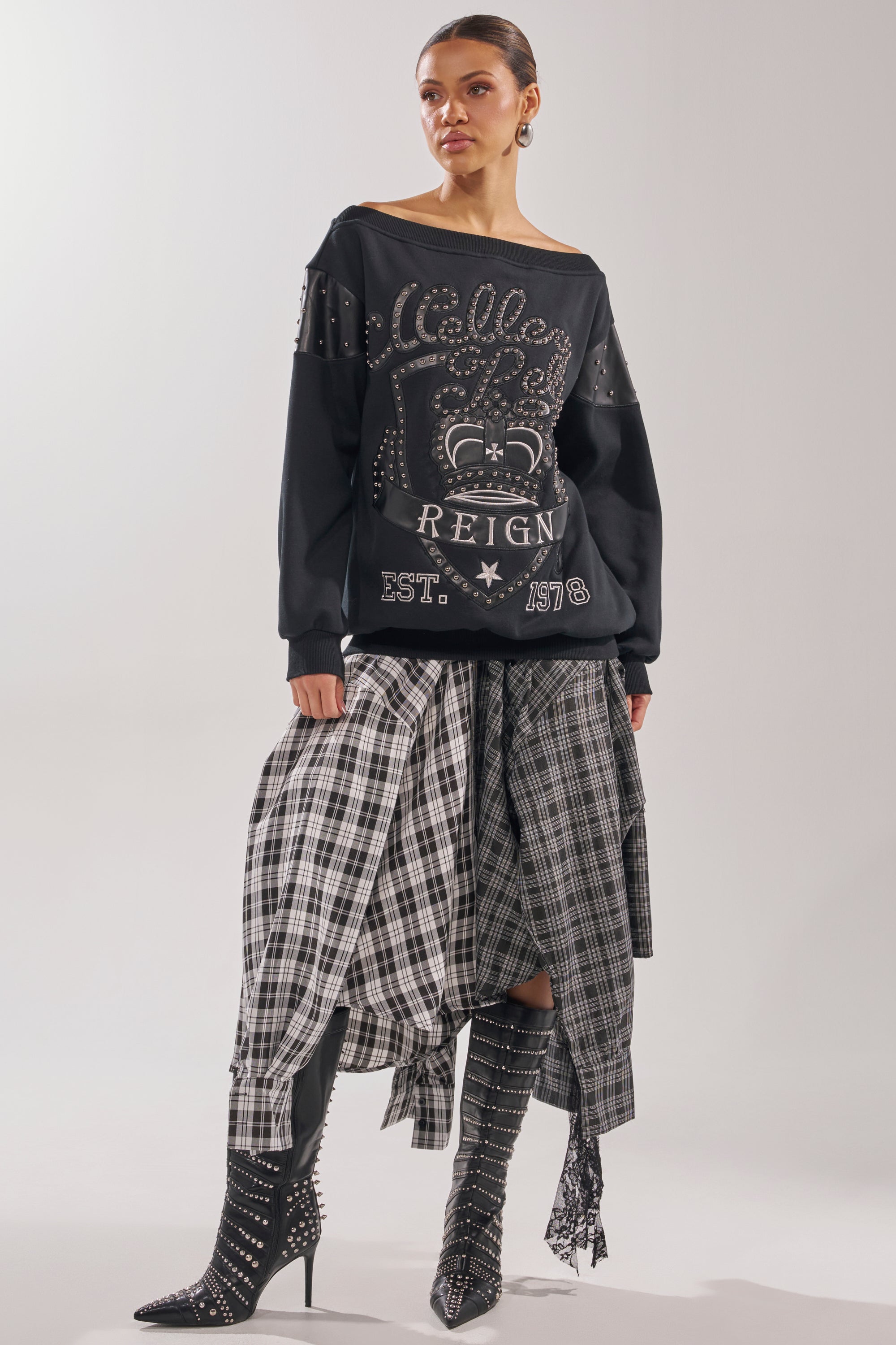 A woman styles the ROYAL OVERSIZED SWEATSHIRT with a layered plaid skirt and studded knee-high boots, her hair tied back and makeup neutral, for an edgy grunge-inspired look. 7