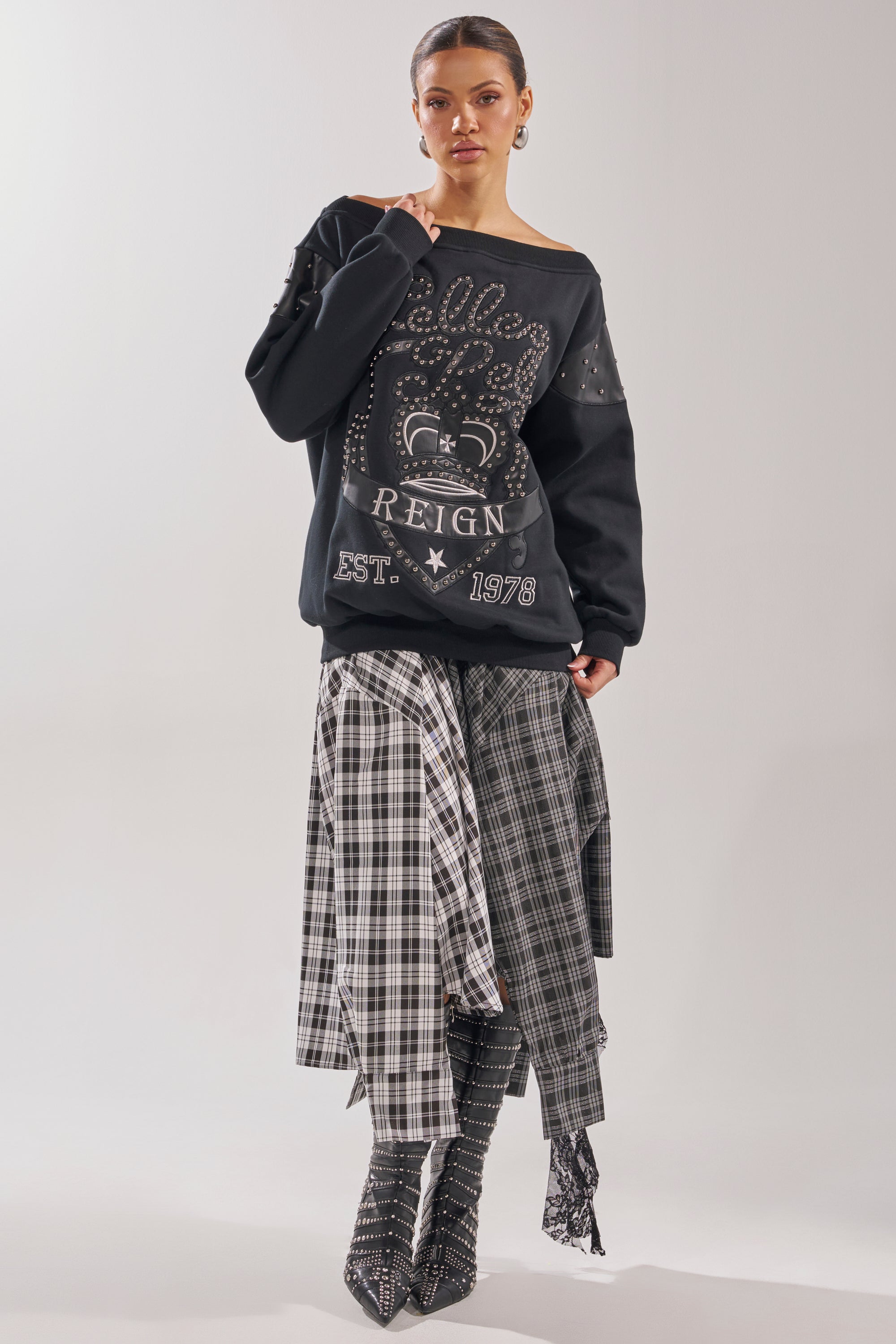 A woman poses against a plain background wearing the ROYAL OVERSIZED SWEATSHIRT, paired with a layered black-and-white plaid skirt and matching plaid boots. 8