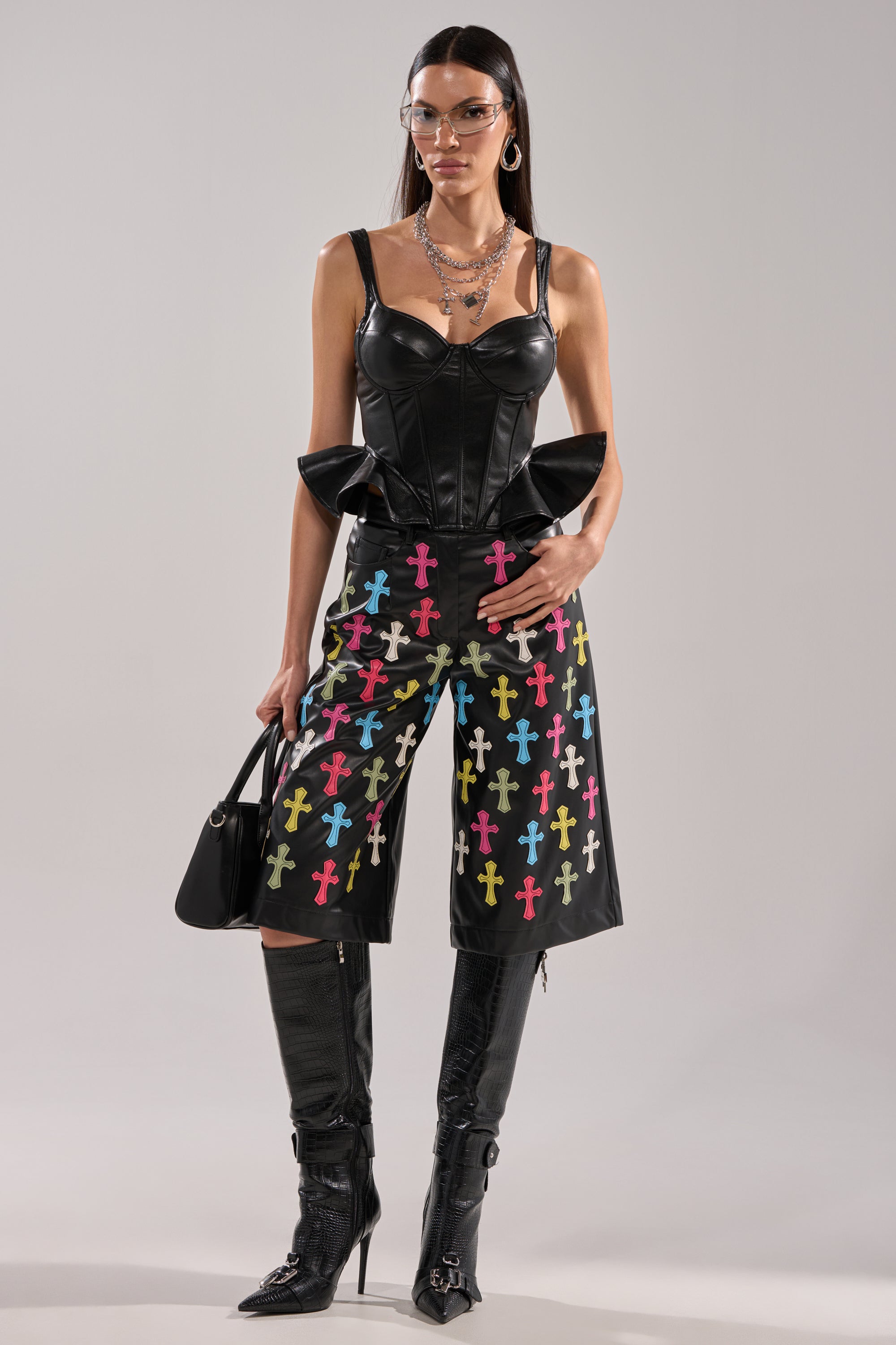 A woman wearing the SEXY AS CAN BE FAUX LEATHER CORSET, colorful cross-patterned black shorts, knee-high black boots, layered necklaces, glasses, and holding a black handbag stands against a plain background. 4