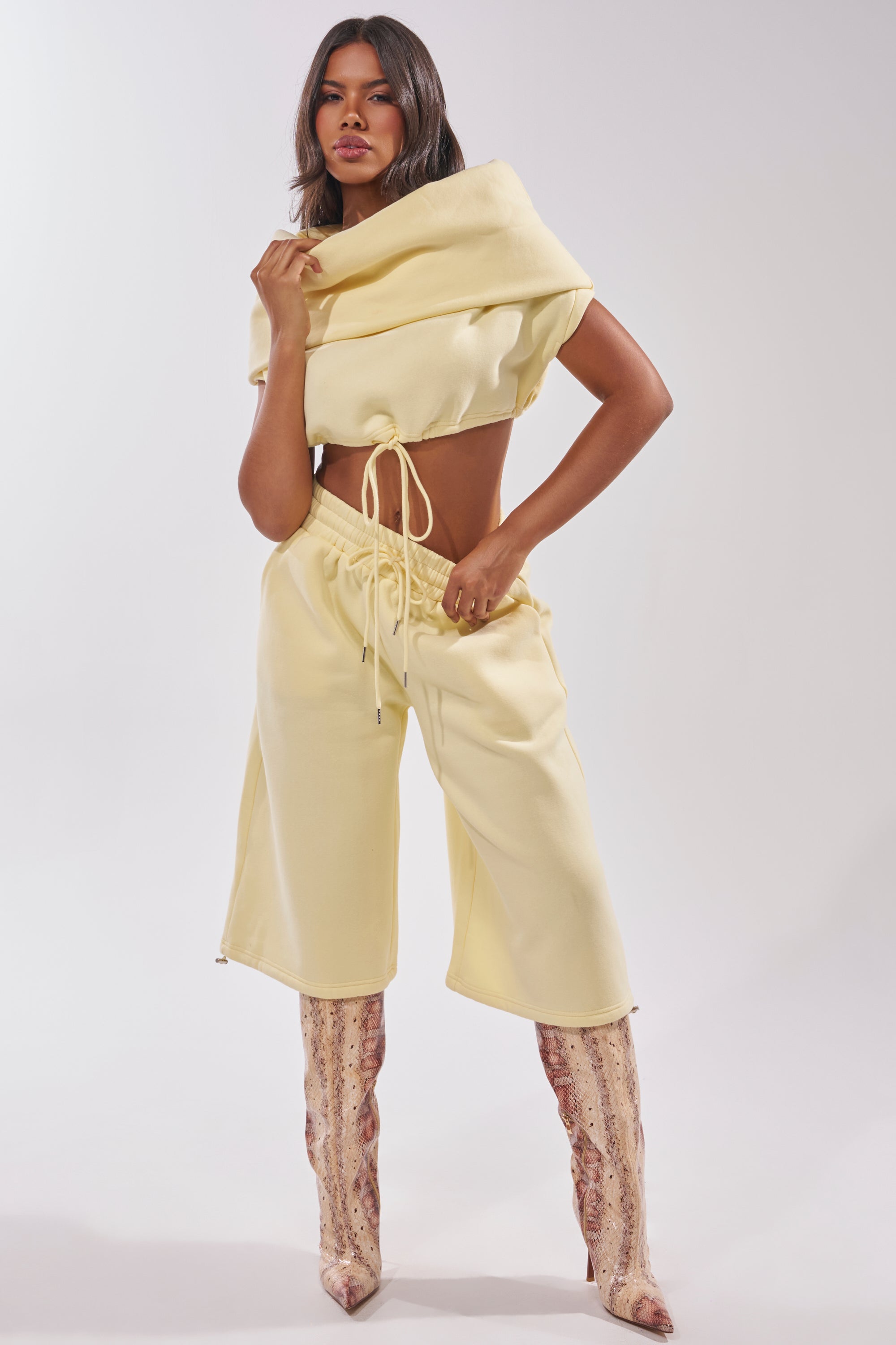 A woman poses confidently in the BABY PLEASE SHORT, a pale yellow, cropped drawstring top paired with matching breathable knit wide-leg, high-waisted pants and knee-high snake print boots against a light background. 0