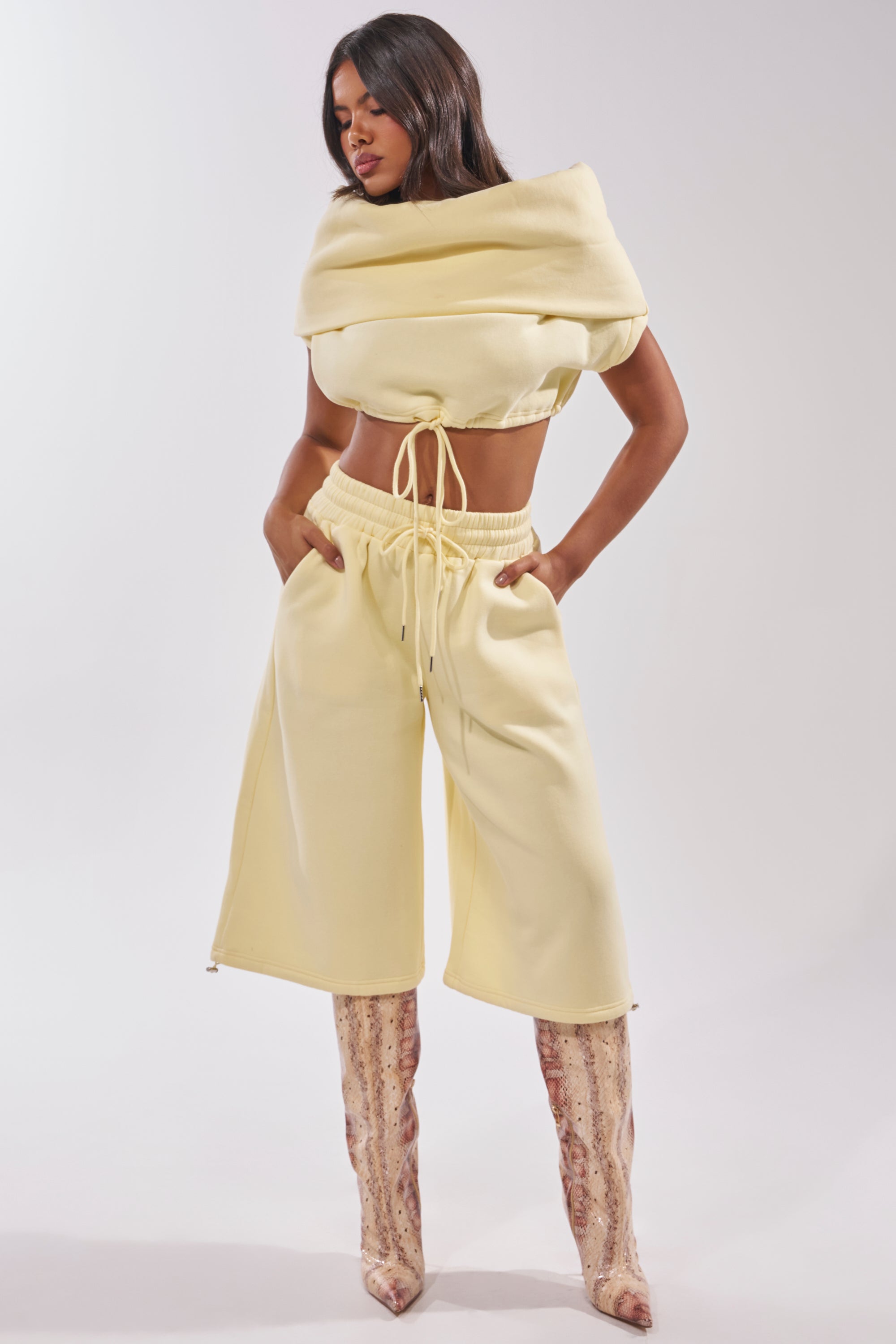 A woman wears the BABY PLEASE SHORT, a pale yellow lightweight knit short with a high-rise elastic waistband, paired with a matching cowl neck top and knee-high snakeskin boots, standing hands-in-pockets against a plain background. 9