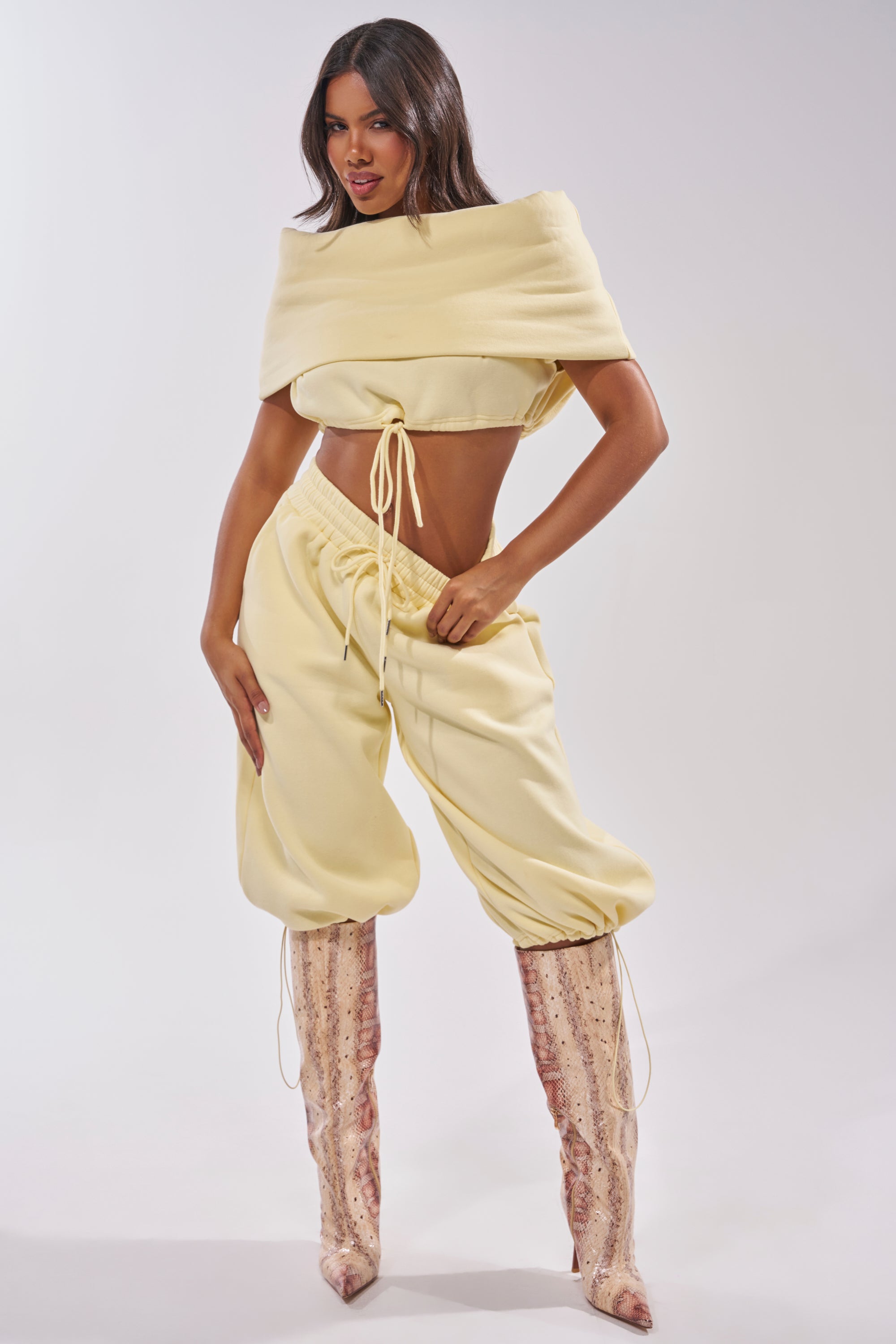 A woman poses confidently in the BABY PLEASE SHORT, a pale yellow cropped top with a draped neckline, paired with high-rise drawstring pants and tall snakeskin boots against a plain light background. 11