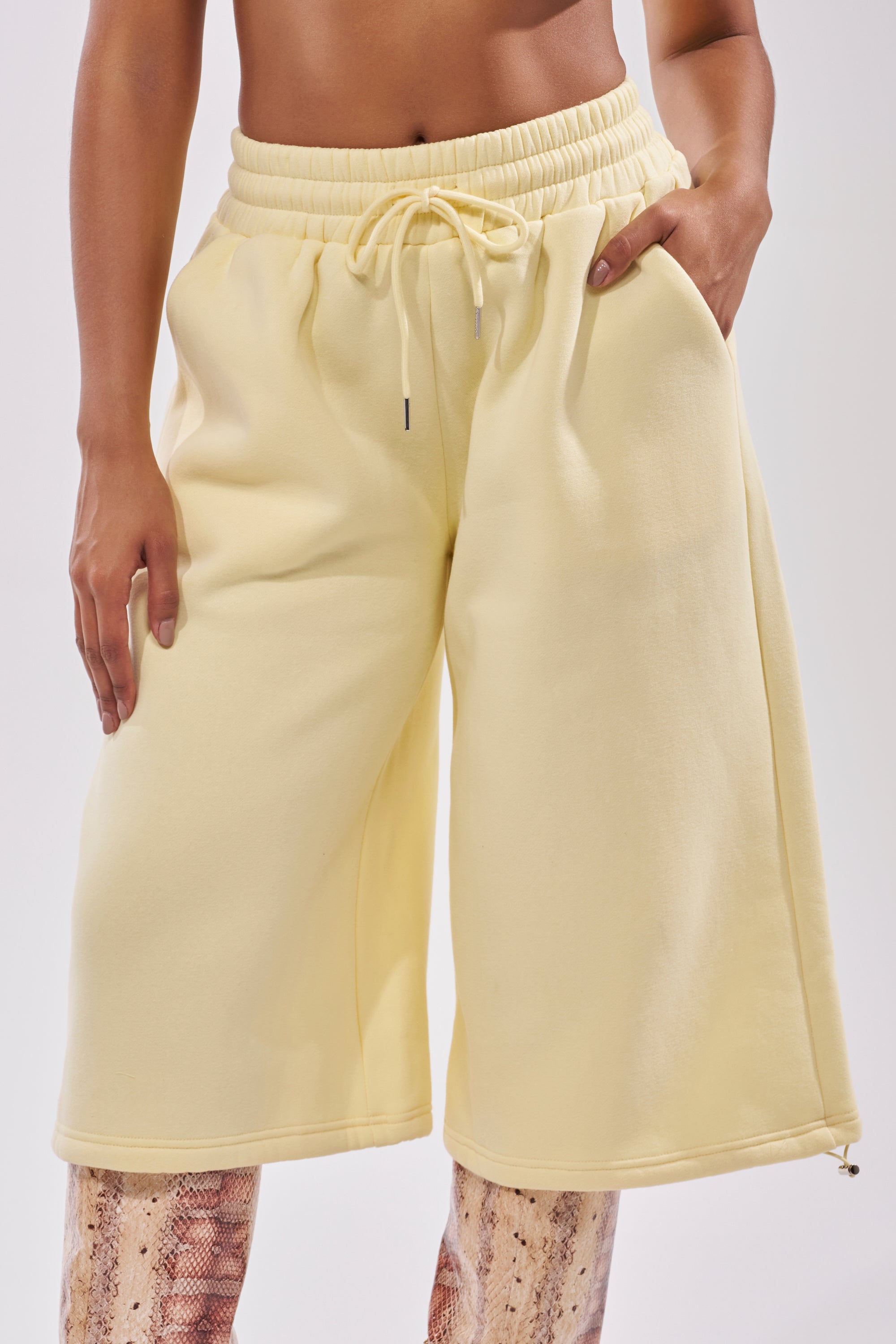 Someone stands with one hand in their pocket, wearing the BABY PLEASE SHORT—pale yellow, lightweight knit lounge shorts with a high-rise elastic waistband—layered over beige and brown snake-print leggings. 12