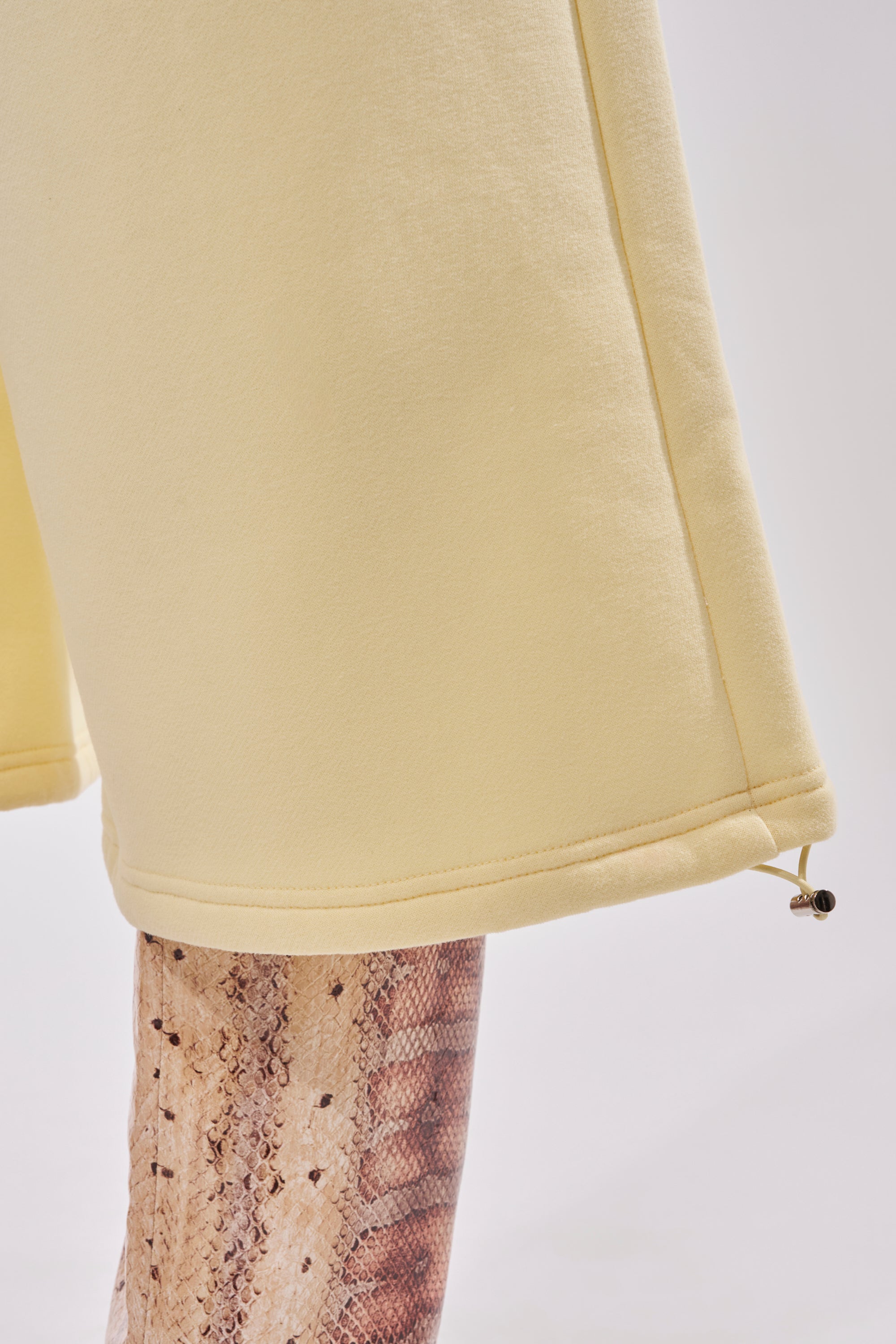 A close-up of a person wearing the BABY PLEASE SHORT in pale yellow, featuring a high-rise elastic waistband and drawstring hem, styled over beige snake-print leggings. 13