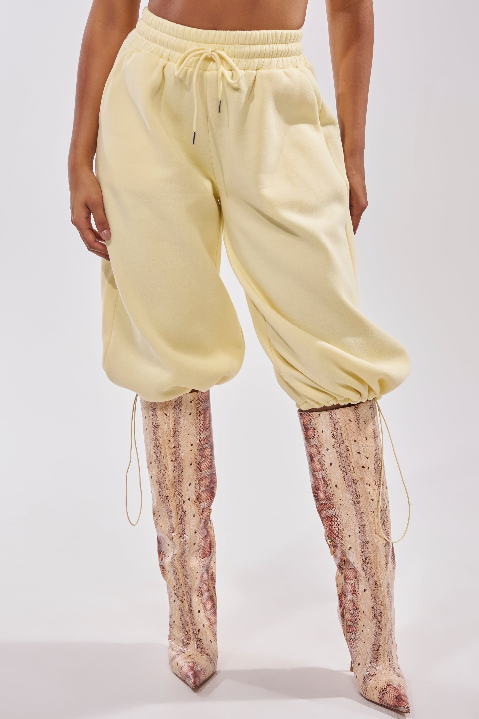 Someone is wearing the BABY PLEASE SHORT, pale yellow jogger shorts with a high-rise elastic waistband and ankle drawstrings, tucked into tall, pointed-toe pink and beige snakeskin boots against a plain white background. 2