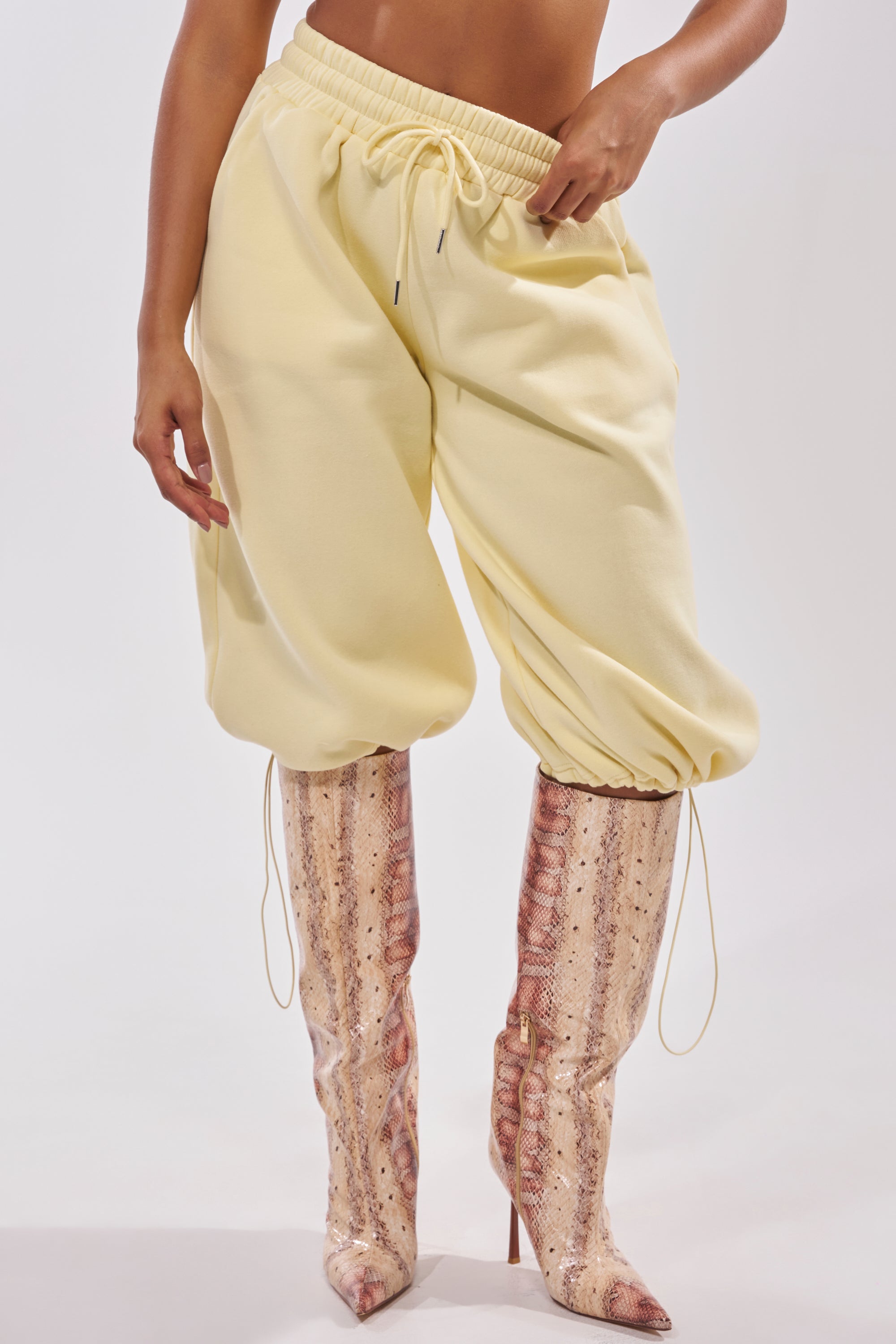 A person wearing the BABY PLEASE SHORT—loose, yellow jogger shorts with a high-rise elastic waistband—tucked into high, pointed-toe snakeskin stiletto boots against a plain, light background. 3