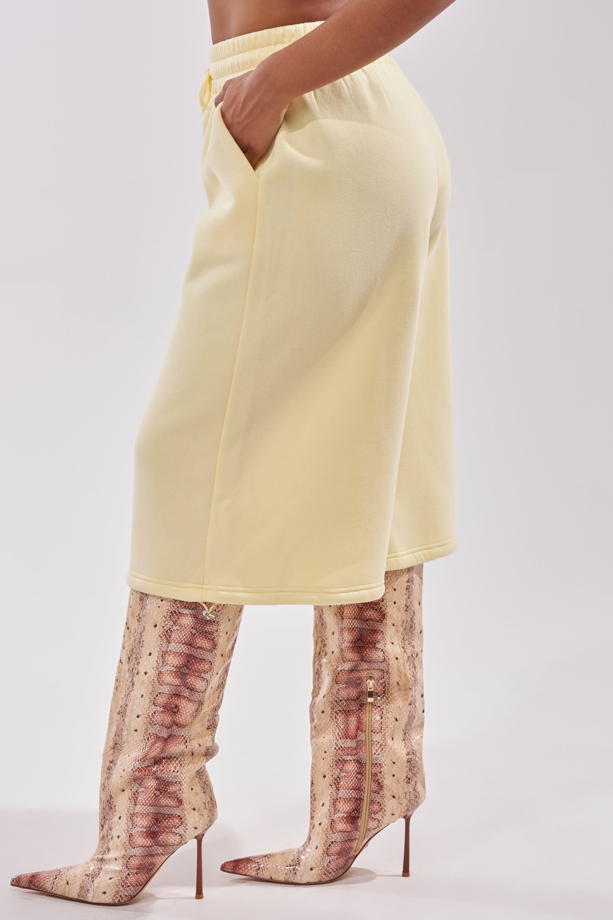 Someone wearing the BABY PLEASE SHORT—pale yellow lounge shorts with a high-rise elastic waistband—paired with tall, brown snakeskin-patterned pointed-toe heeled boots, stands with one hand in their pocket against a plain background. 4
