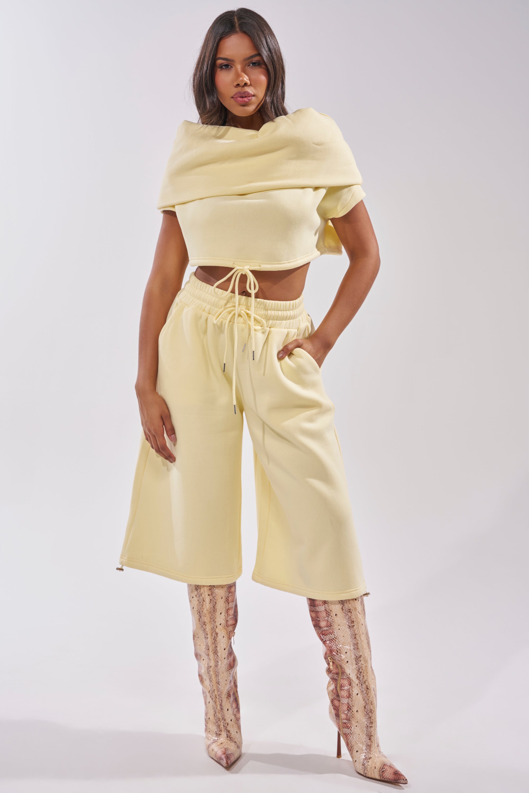 A woman stands confidently in the BABY PLEASE SHORT, styled with pink snakeskin knee-high boots. She has long dark hair and poses with one hand on her hip against a plain background. 6