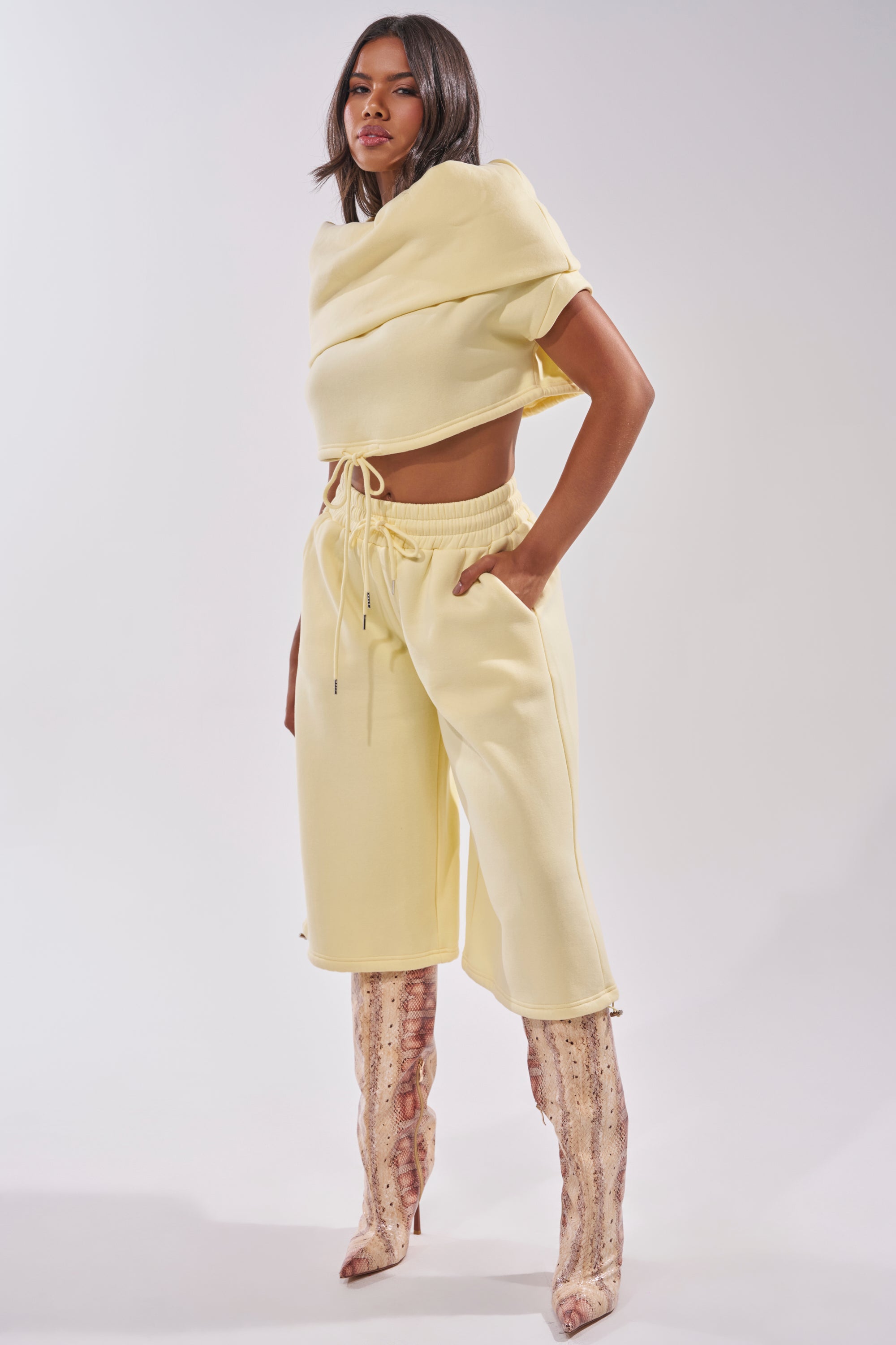 A woman stands confidently in the BABY PLEASE SHORT—lightweight, high-rise yellow lounge shorts paired with a pale yellow cropped hoodie and knee-high snakeskin boots against a plain light background. 8