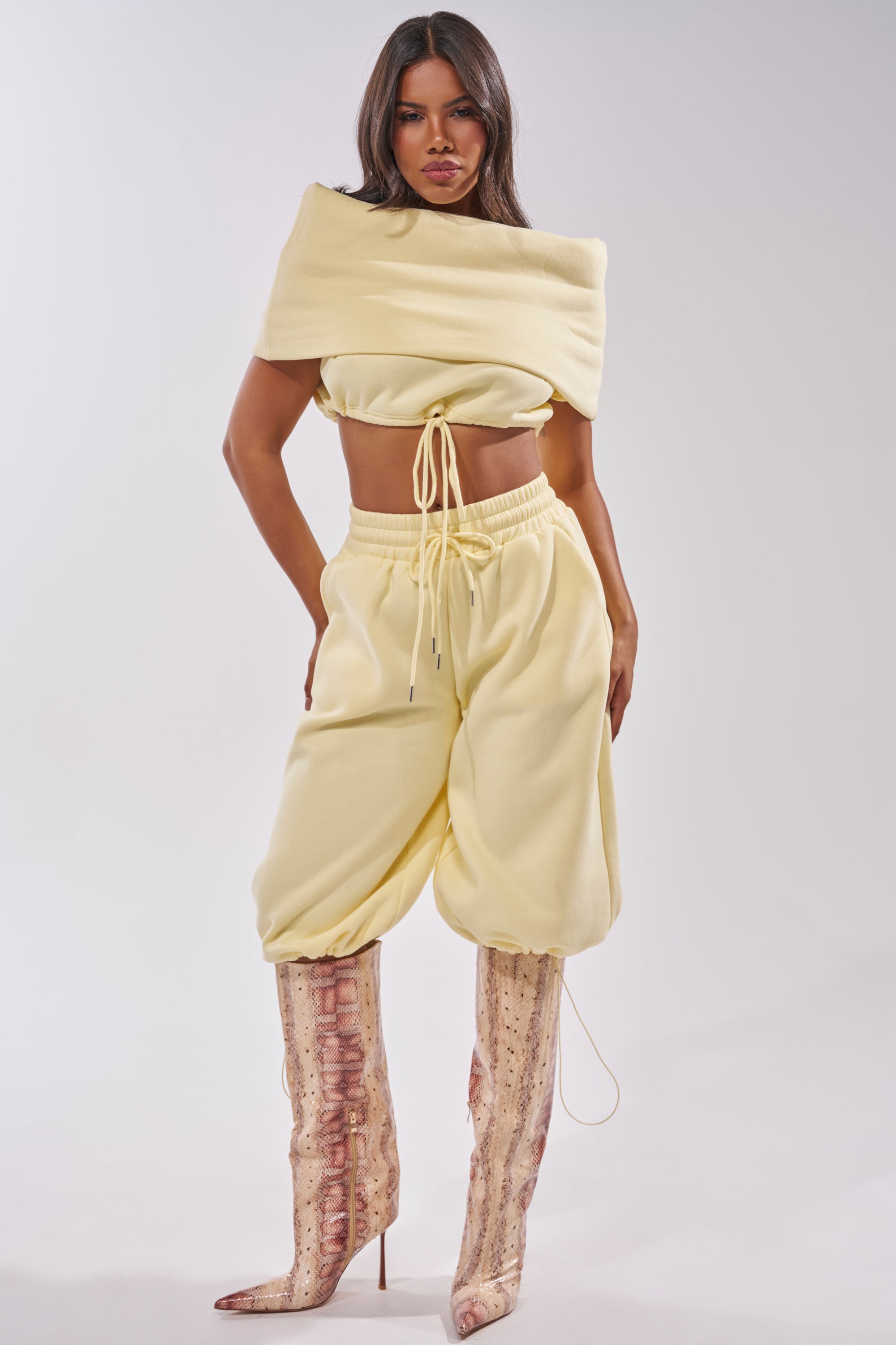 A woman faces forward in the BABY PLEASE SWEATSHIRT, a baby yellow cropped lounge top with matching baggy pants and tall, textured heeled boots against a seamless light gray background. 5