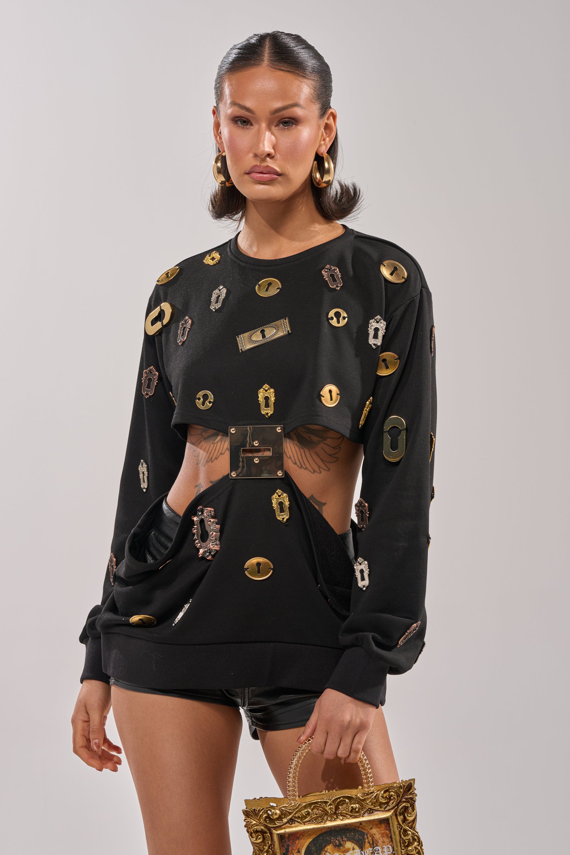 A woman exudes confidence in the KEY CHAIN TOP with metallic accents, paired with black shorts, hoop earrings, and a baroque-style gold-framed handbag. Her slicked-back hair completes this bold streetwear look. 1