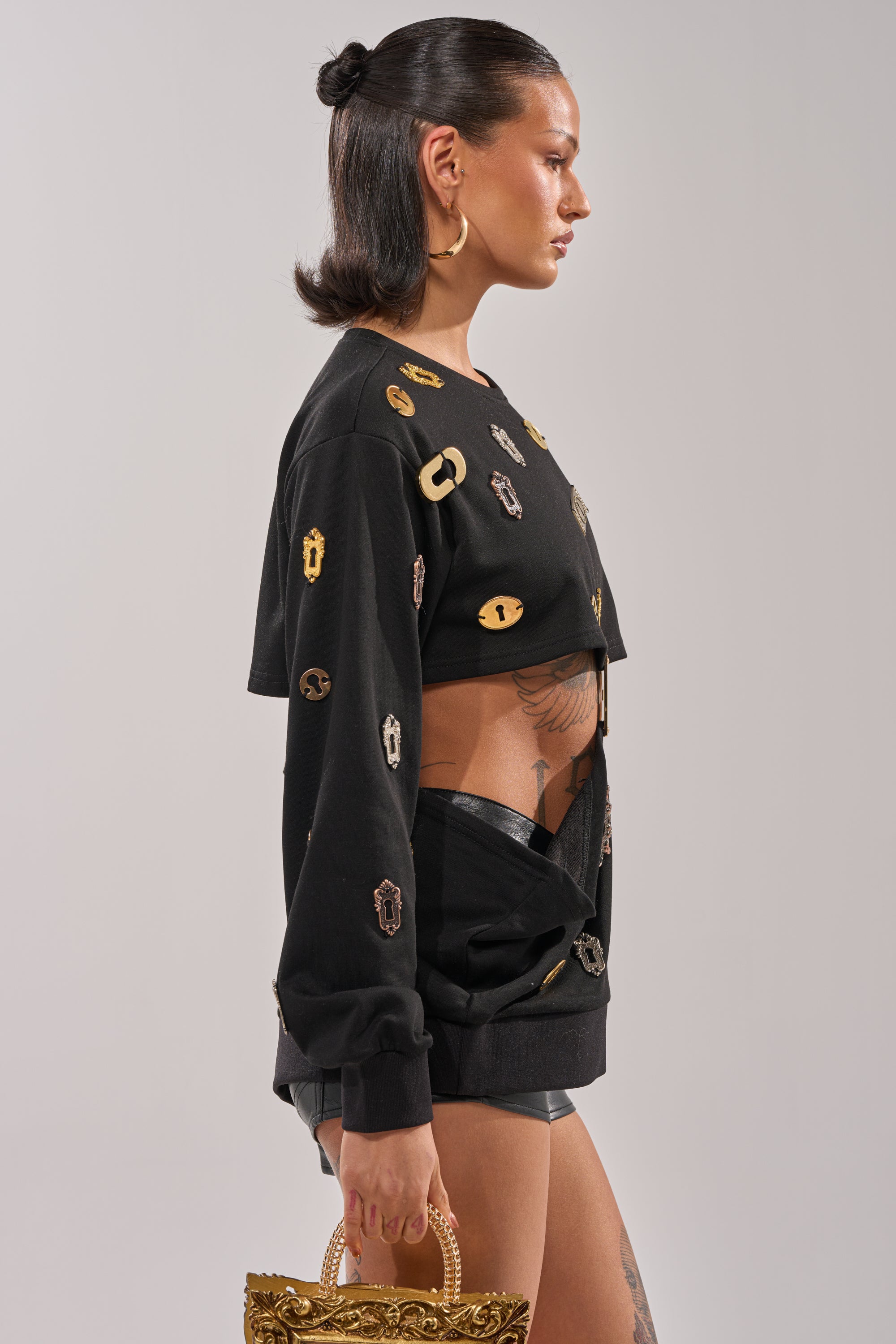A woman with sleek dark hair wears the KEY CHAIN TOP—a black cutout top with metallic details—paired with black shorts, gold hoop earrings, and an ornate gold handbag. Tattoos on her torso and thigh add to her edgy streetwear style. 2