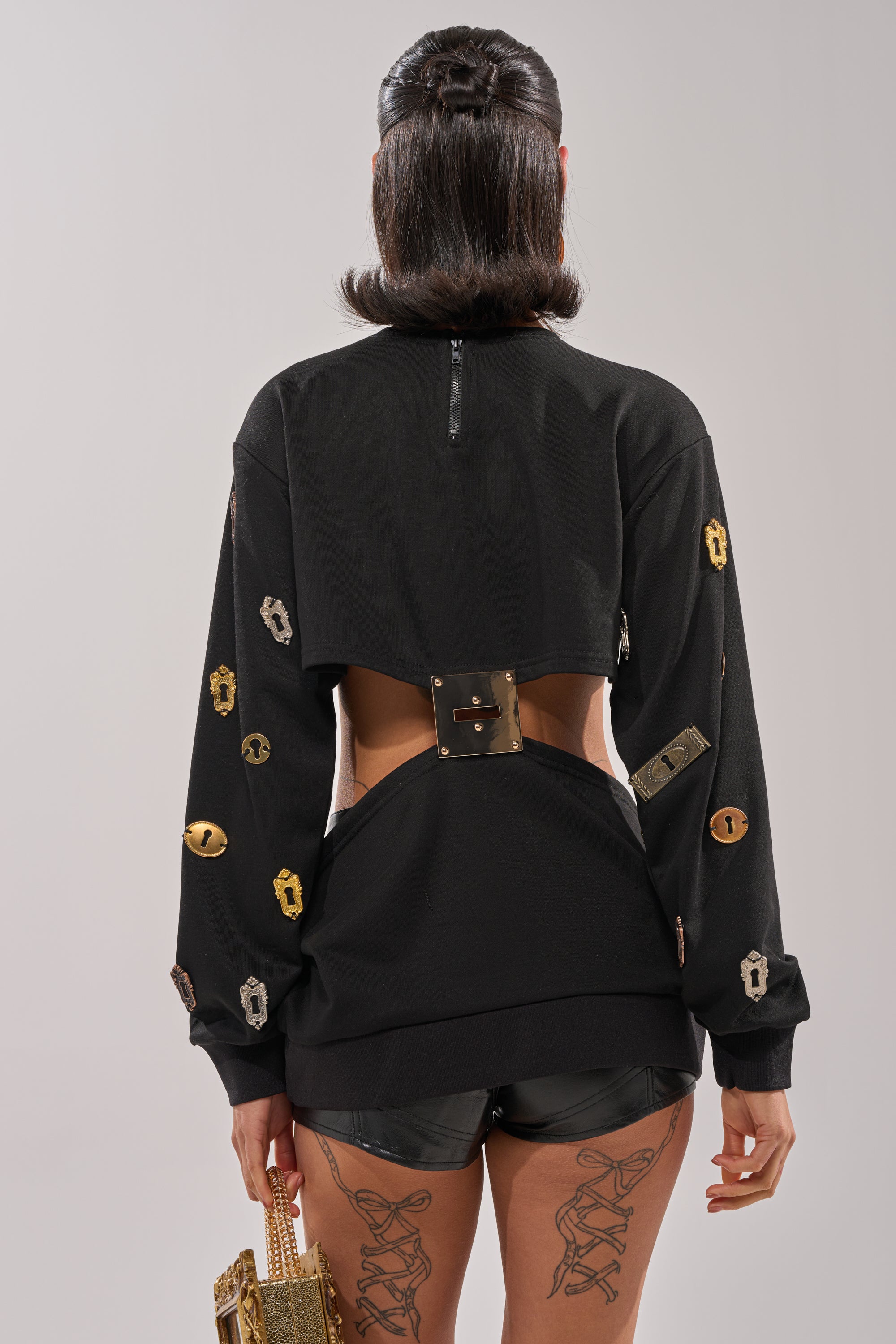 A woman with dark, shoulder-length hair stands facing away in the KEY CHAIN TOP—a black cutout design with metallic accents—paired with black shorts, a gold bag, and lace-up tattoo details on her thighs for bold streetwear style. 3