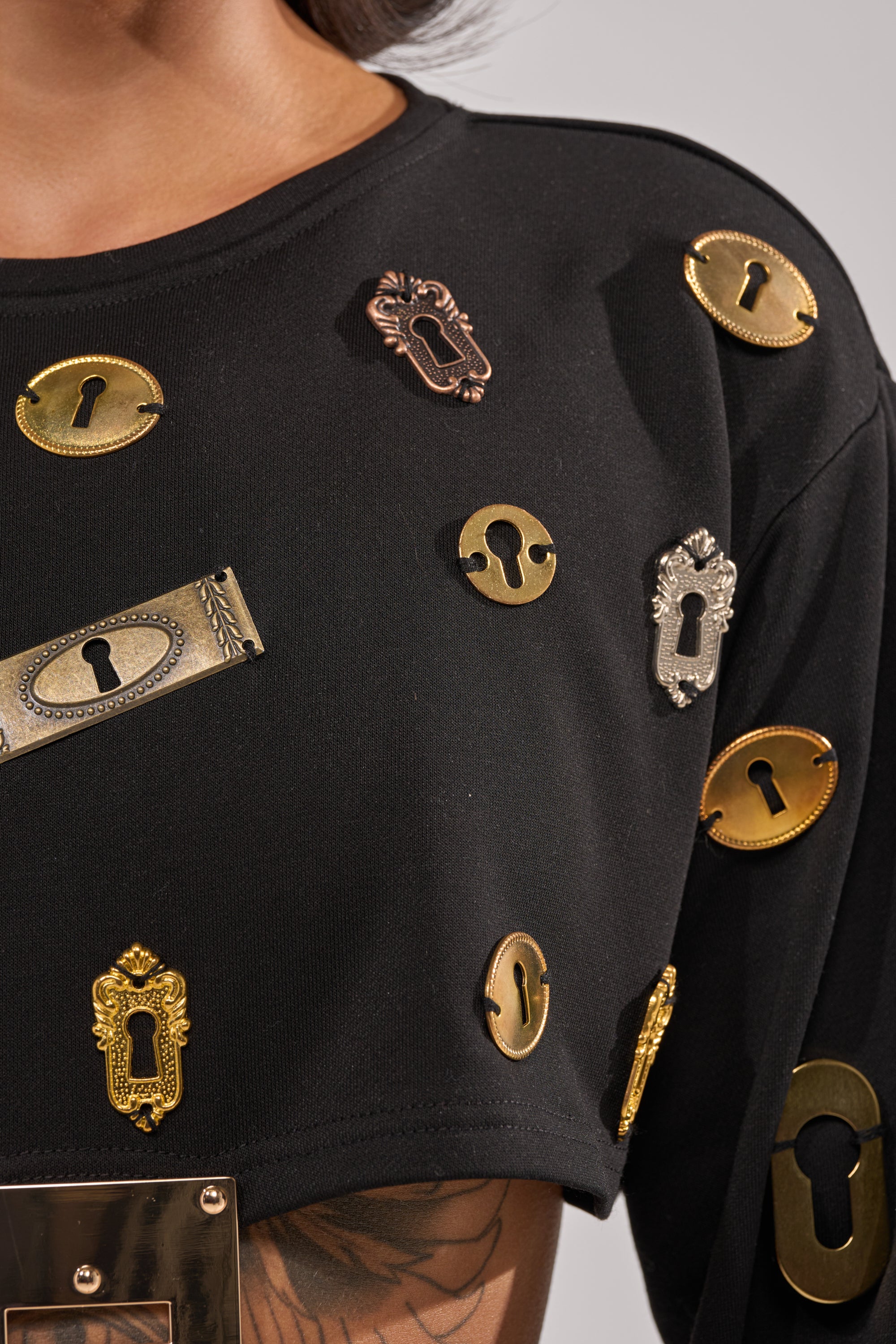 A close-up of the KEY CHAIN TOP, a streetwear black cutout top featuring gold and bronze keyhole plates arranged across the fabric as metallic embellishments. 7