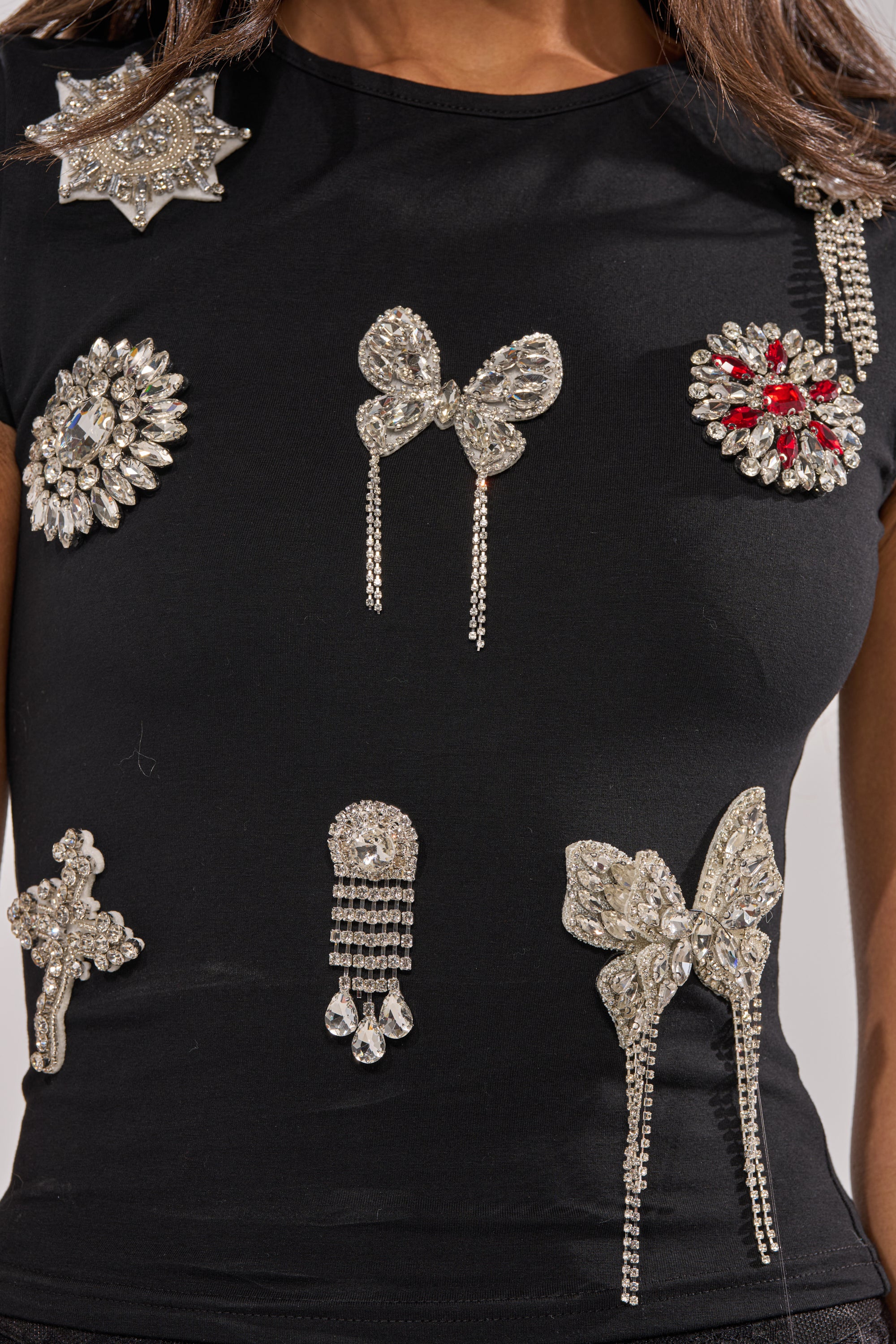 A person wears the ORNAMENTAL RHINESTONE TEE, a black stretch-knit crew neck adorned with sparkling rhinestone brooches in bow, flower, and abstract shapes for a glamorous embellished look. 6