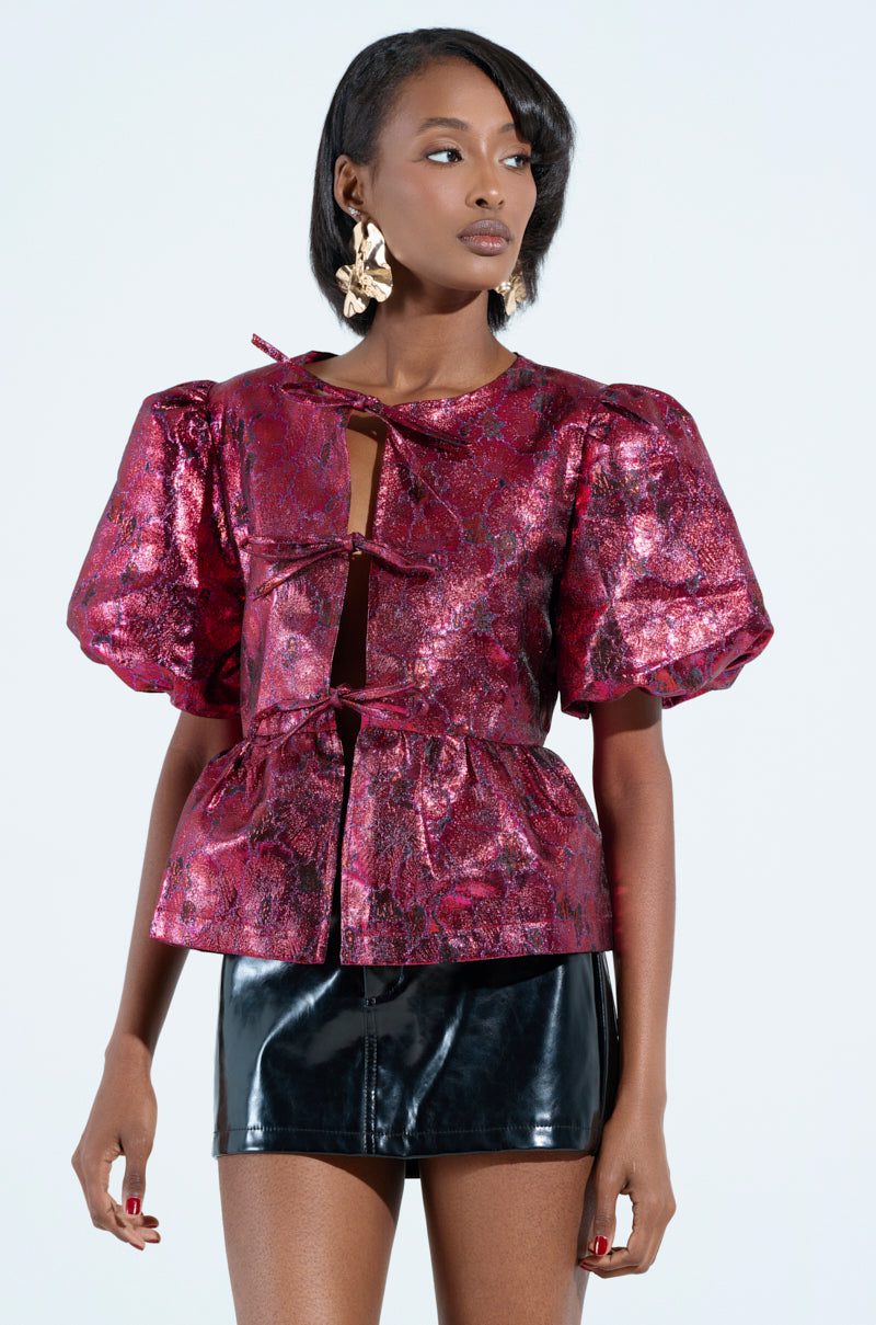 A woman poses against a light blue backdrop, her long hair flowing past large earrings. She's wearing the shimmering "ITS GOES LIKE BLOUSE" with romantic puff sleeves and decorative bows, paired perfectly with a black leather mini skirt.