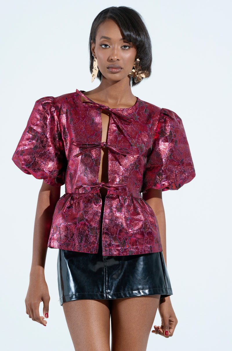A person wearing the "ITS GOES LIKE BLOUSE," a shimmering brocade top with romantic puff sleeves and a front bow, paired with a black leather mini skirt. They have sleek hair and pose against a plain light background.