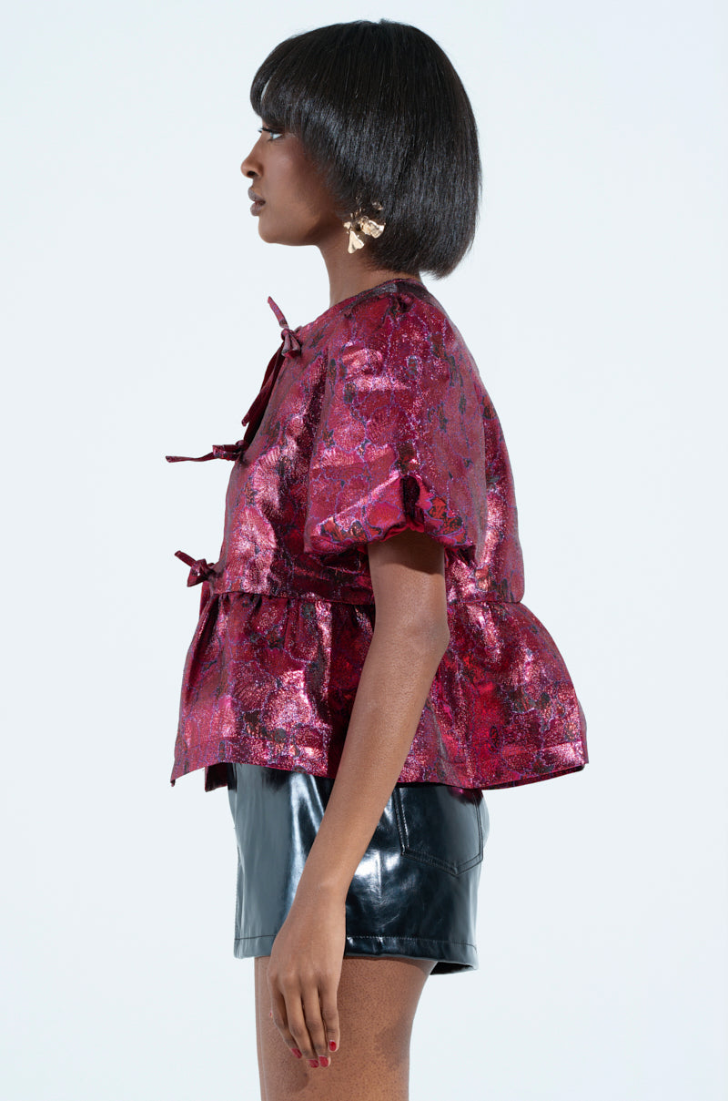 With a bob haircut, the person poses in profile, wearing the shimmering "ITS GOES LIKE BLOUSE," featuring bows and romantic puff sleeves, paired with a black leather skirt against a light-colored background.