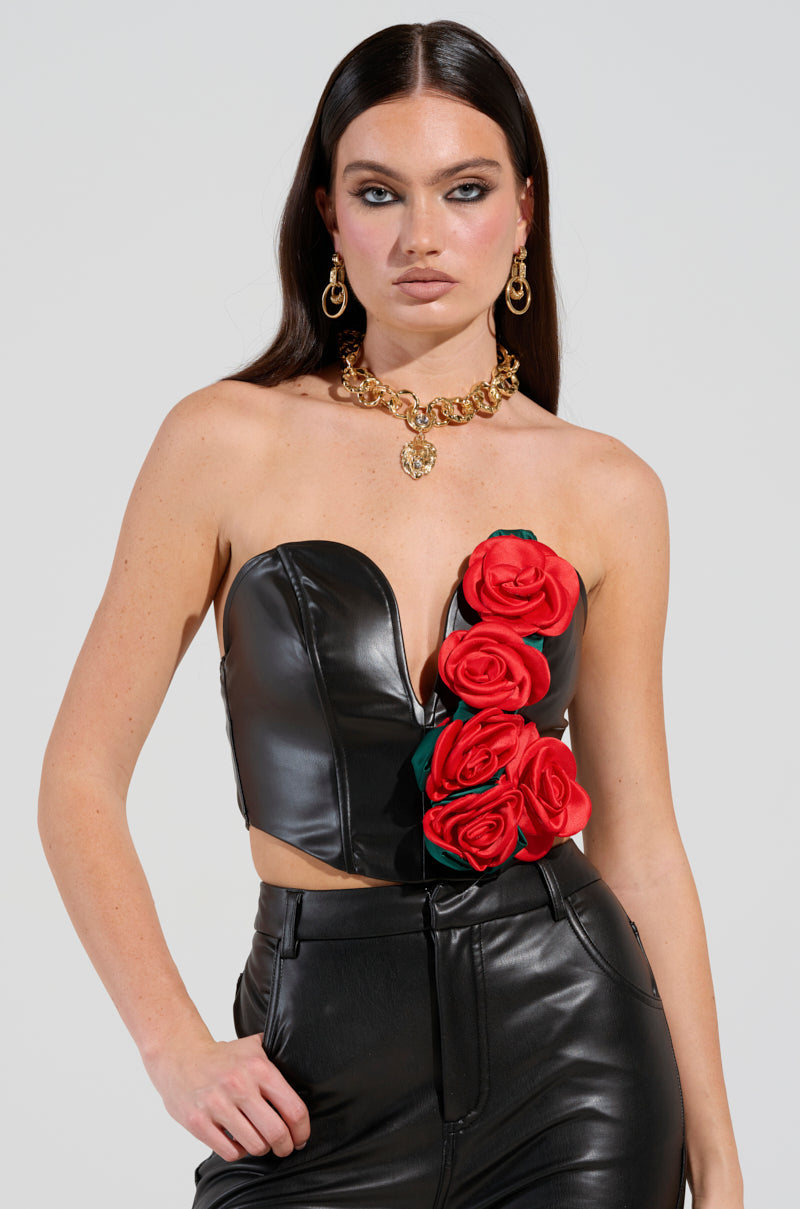 A woman stands confidently in the ROSE PETALS CORSET TOP IN BLACK MULTI, featuring 3D satin rose appliques, paired with sleek black leather pants. She accessorizes with gold hoop earrings and a chunky gold necklace. Her long dark hair falls straight, complementing her bold makeup and striking eyes.