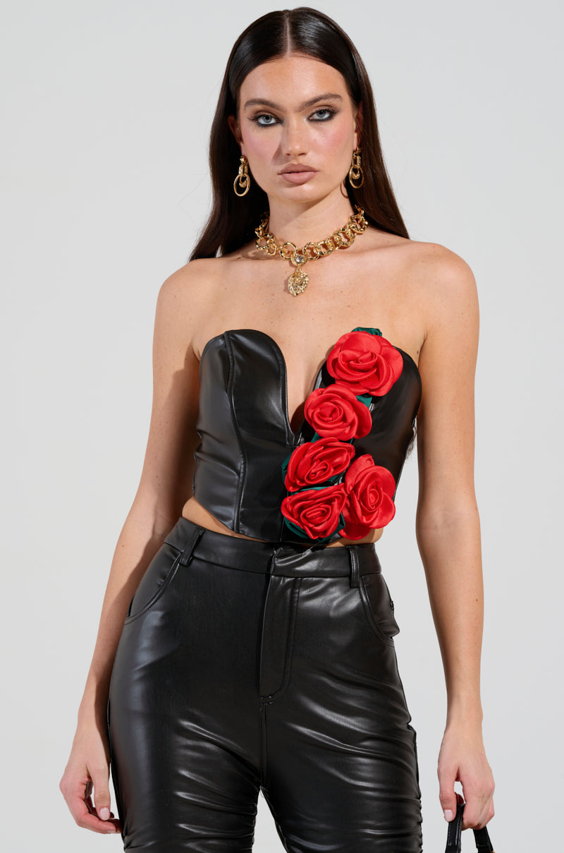 A woman with long dark hair wears the ROSE PETALS CORSET TOP IN BLACK MULTI, a strapless black faux leather piece embellished with 3D satin rose appliques. She pairs it elegantly with black leather pants and accessorizes with gold hoop earrings and a gold necklace showcasing a lion's head pendant, radiating elegance and style.