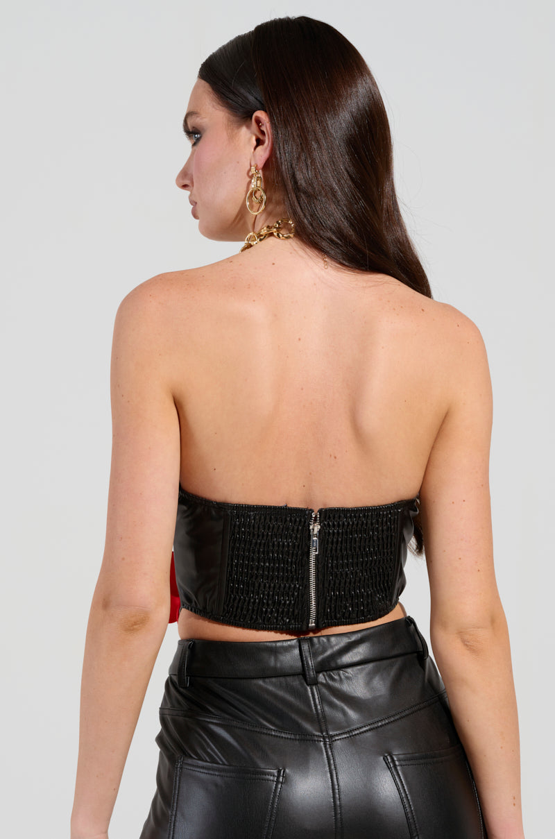 A woman with long dark hair faces away, wearing the ROSE PETALS CORSET TOP IN BLACK MULTI, made of strapless faux leather. Her outfit is completed with matching pants and features a zipper at the back. She accessorizes with large hoop earrings and a gold necklace, adding a touch of elegance reminiscent of 3D satin rose appliques against the plain gray background.