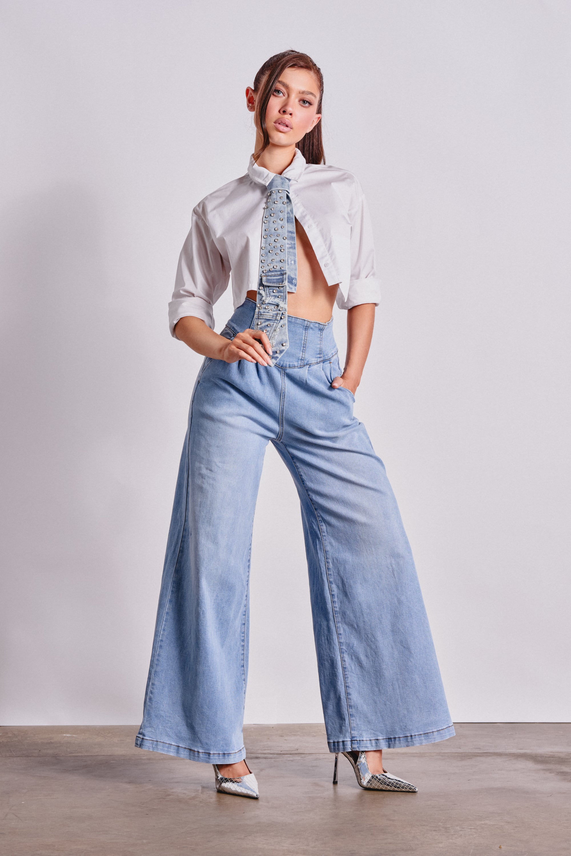 A woman poses confidently in the JENNY WIDE LEG DENIM jeans, paired with a white cropped button-up shirt, patterned necktie, and pointed-toe heels against a plain white background. 0