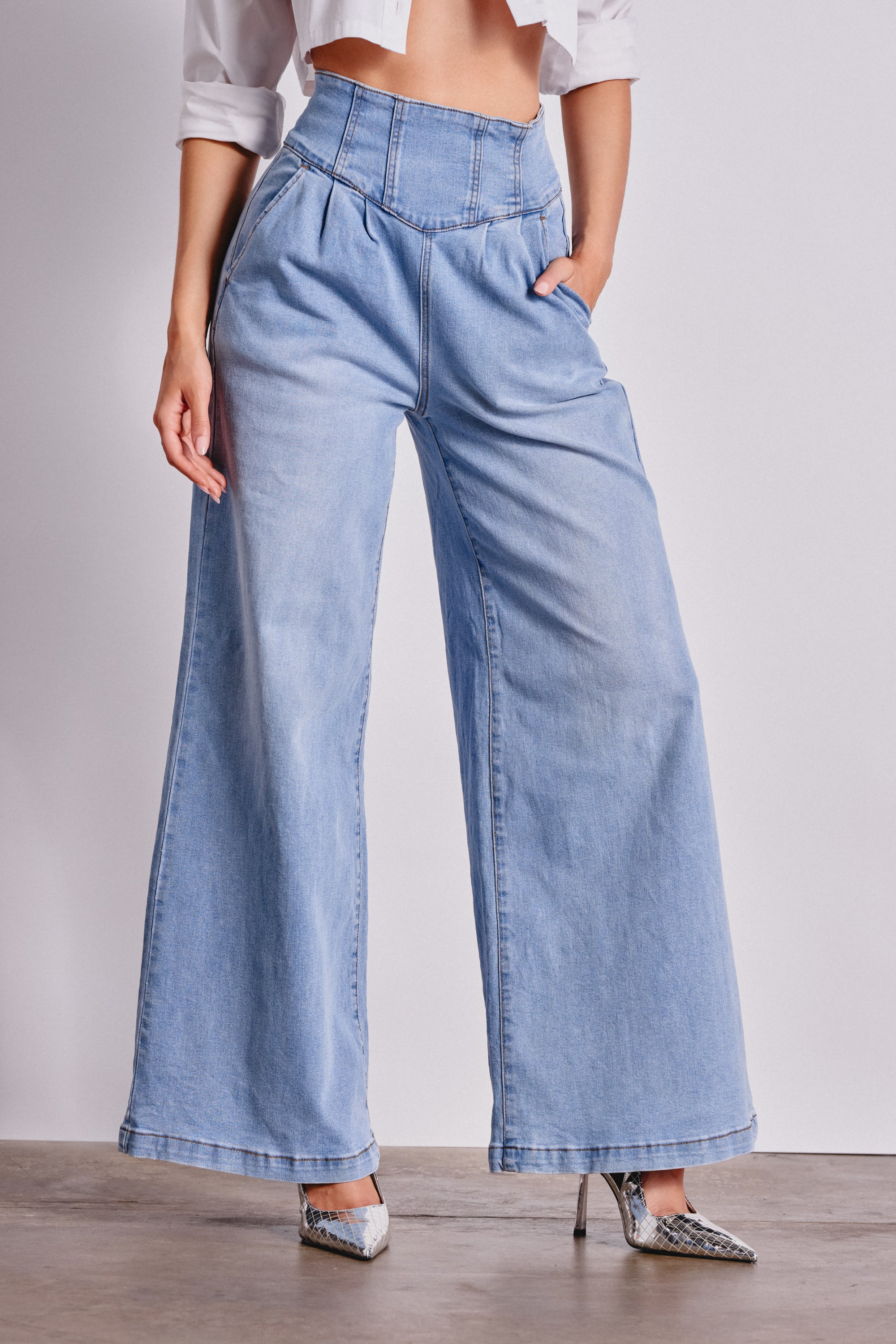 A person in JENNY WIDE LEG DENIM in light blue, a cropped white shirt, and snakeskin-patterned heels stands with one hand in their pocket against a plain background. 1