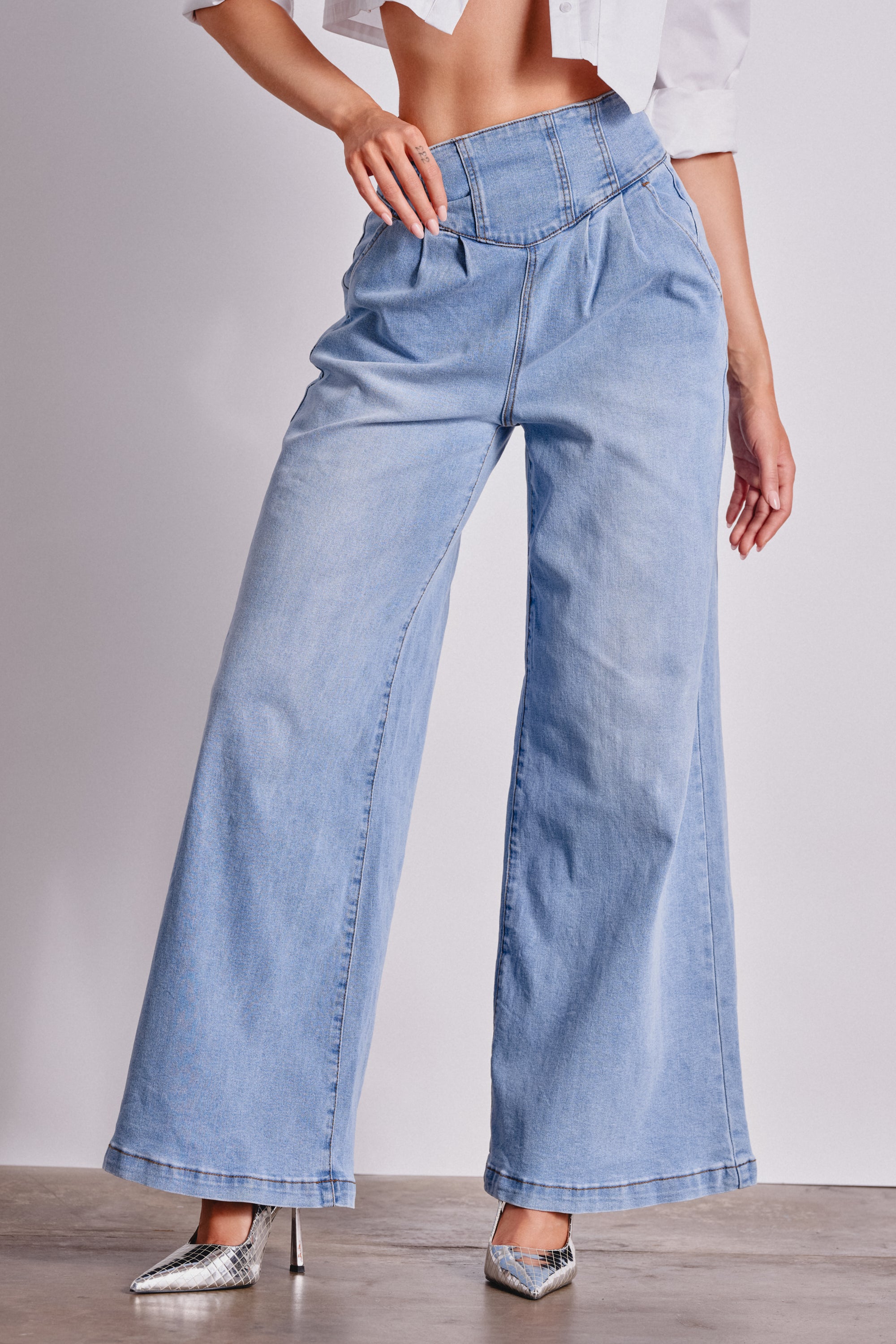 A model wears the JENNY WIDE LEG DENIM, featuring a high-rise fit, sculpted waistband, and pleats, styled with a white cropped shirt and silver pointed-toe heels against a plain neutral background. 2