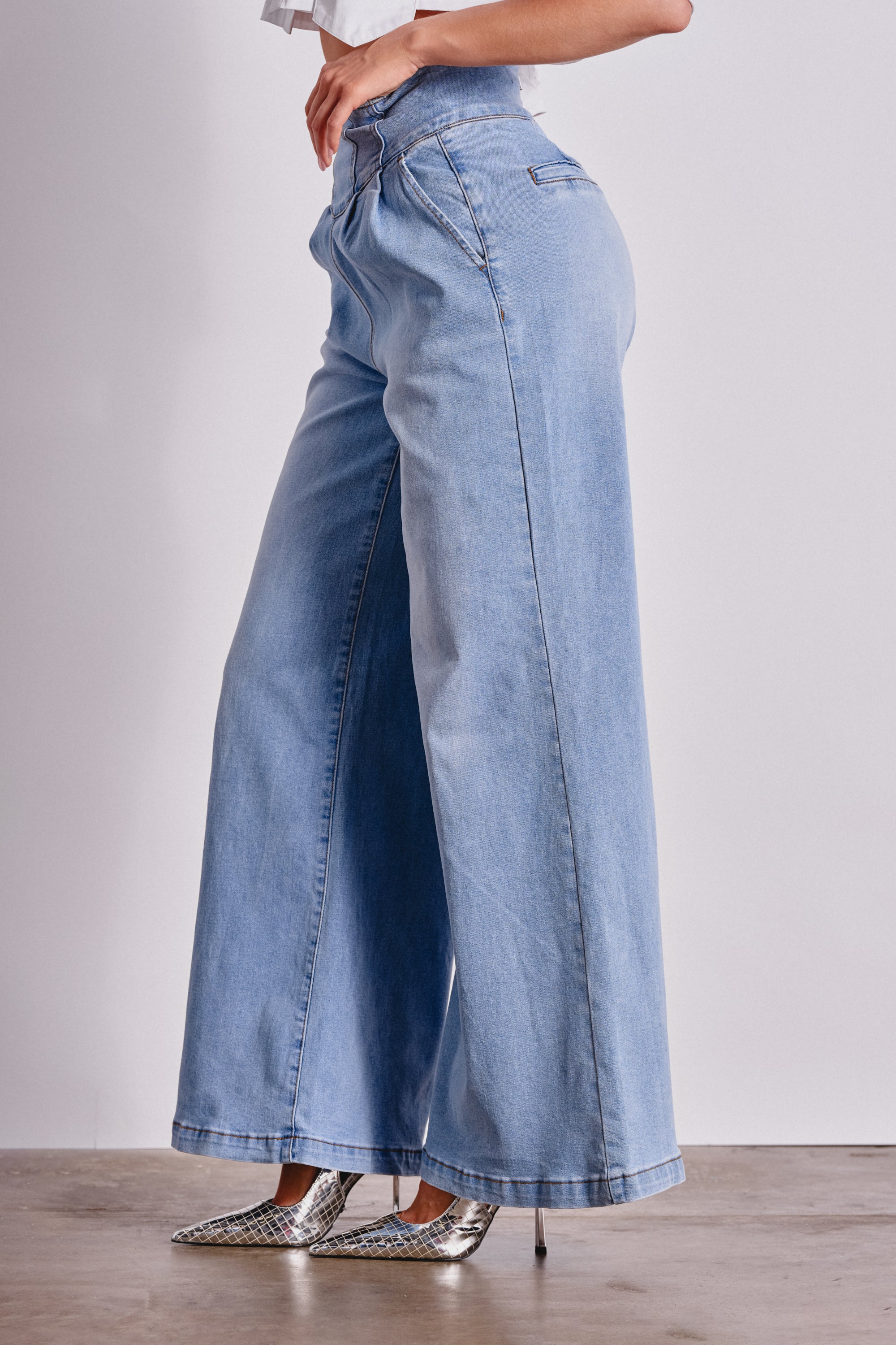 A person in JENNY WIDE LEG DENIM—light blue high-rise jeans with a sculpted waistband and wide legs—pairs them with pointed silver heels, posing sideways against a plain backdrop. 3