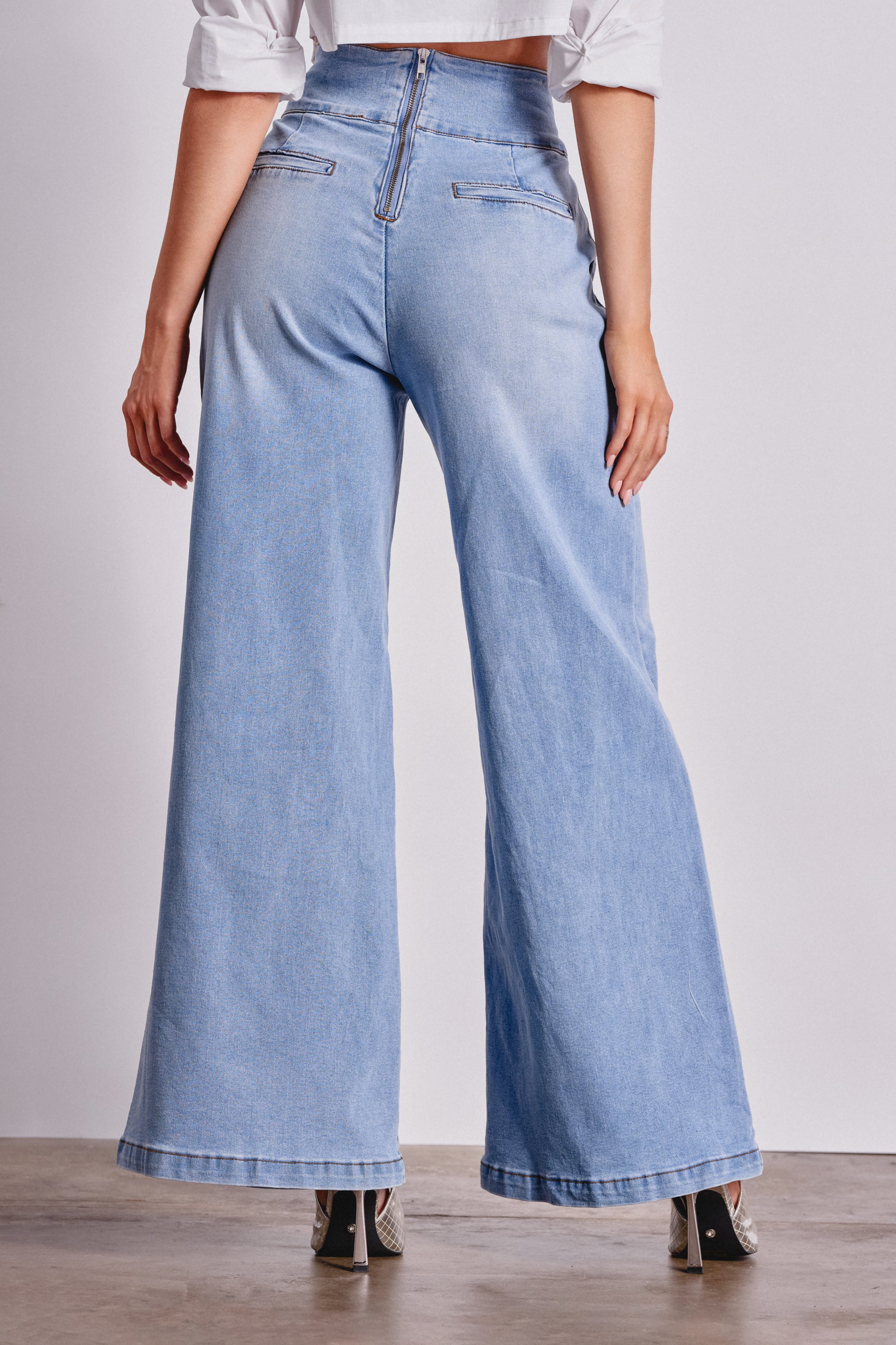 A person stands on a light floor wearing JENNY WIDE LEG DENIM—light blue, high-waisted wide-leg jeans with a sculpted waistband and back pockets—paired stylishly with heeled shoes, hands resting at their sides. 4