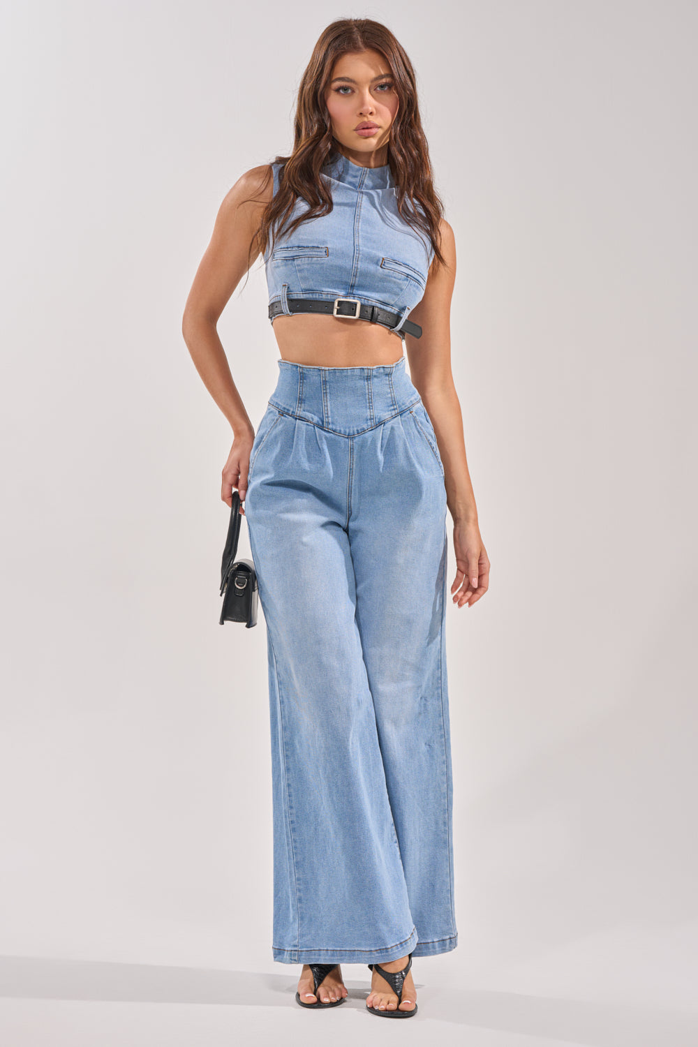 A woman stands against a plain background in streetwear style, wearing the sleeveless, high-neck belted JENNY DENIM BLOUSE with high-waisted wide-leg denim pants, black strappy heels, and holding a small black handbag. 9